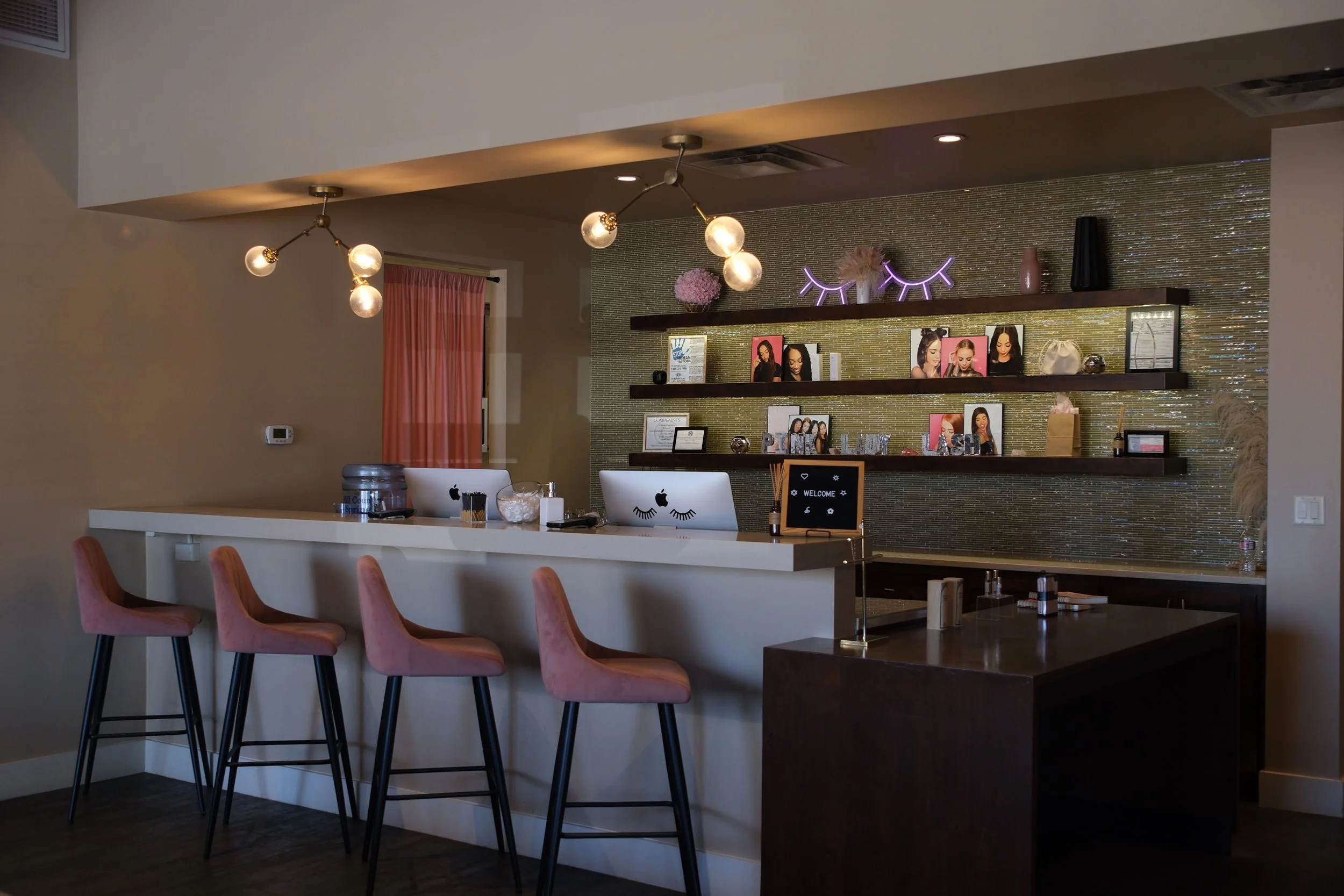 A modern salon reception area with a white counter, four pink velvet chairs, and a wall with shelves displaying hair and beauty products, framed photos, and decor including a neon eyelash sign.