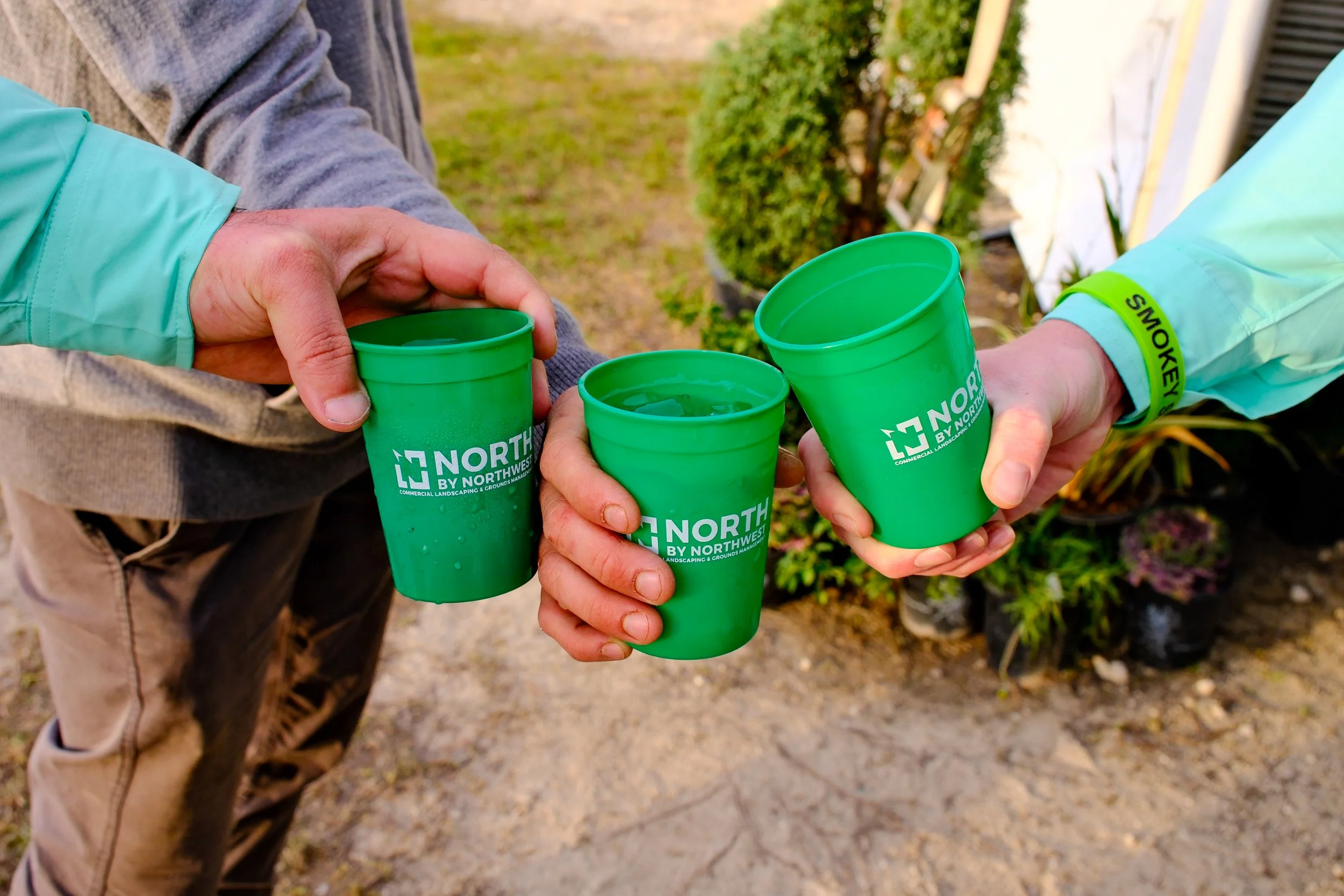 Three people clinking green cups with water, outdoors with plants in the background.