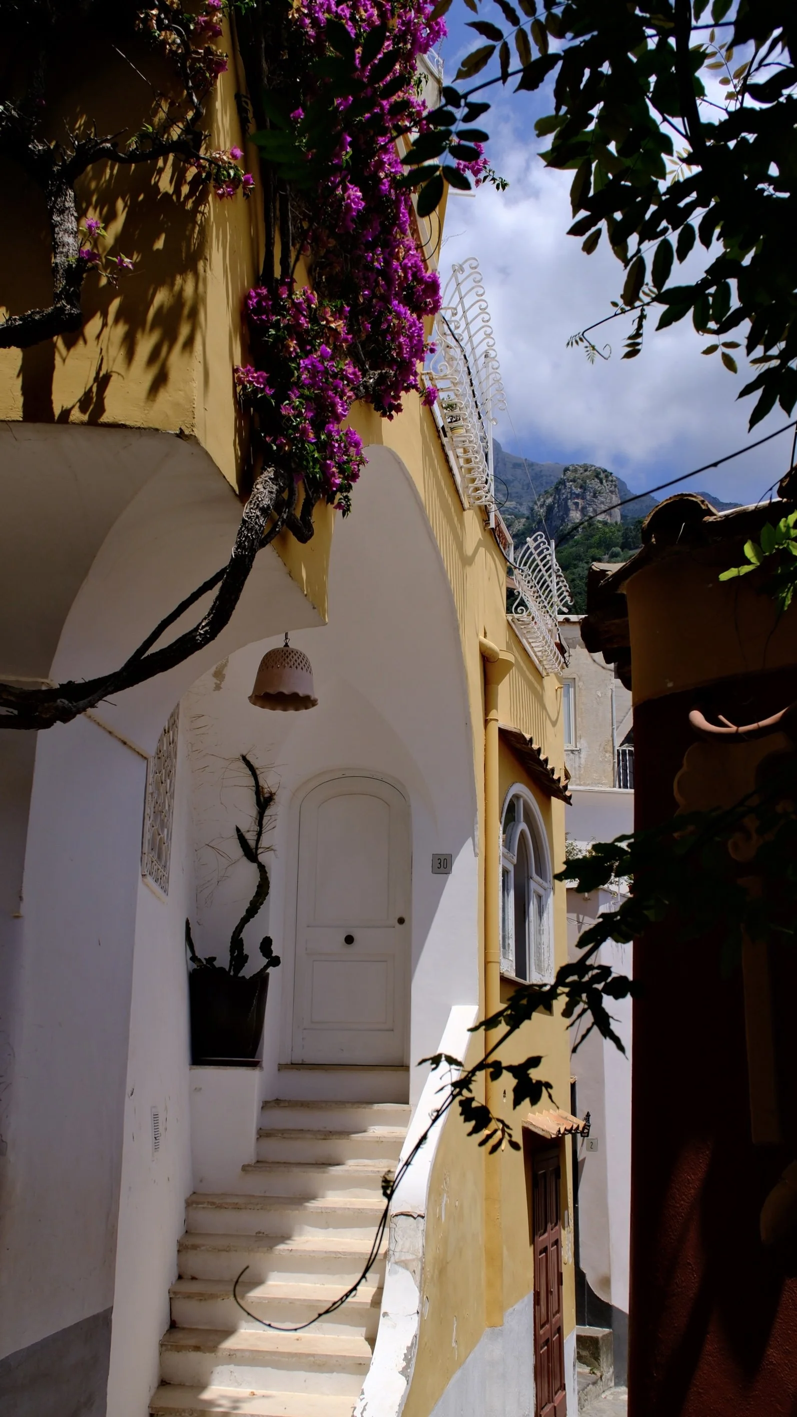 A narrow alleyway leading to a white arched doorway with steps, surrounded by colorful buildings, flowering plants, and mountain views in the background.