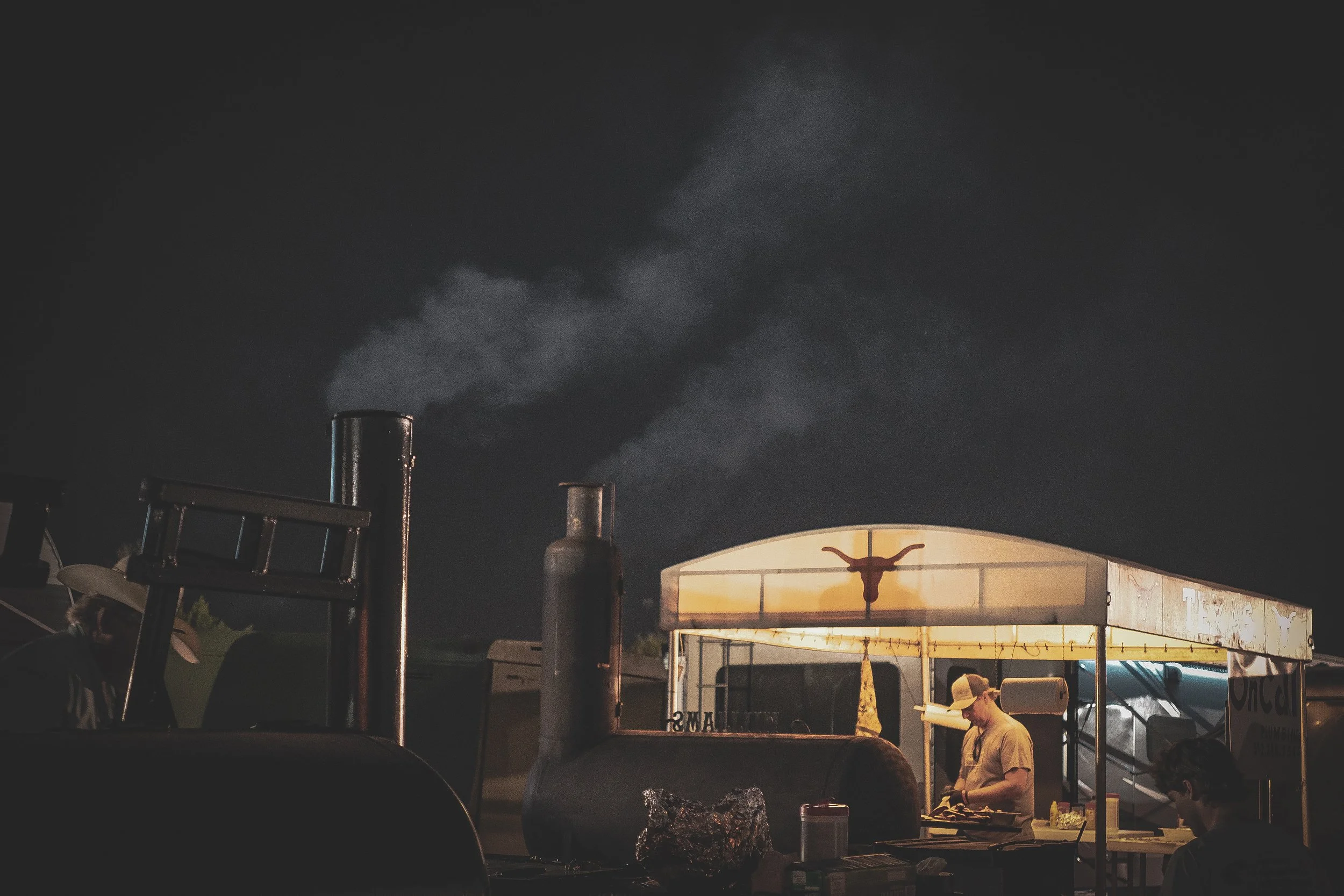 Night scene at a food stand with a man preparing food, illuminated by warm light, smoke rising from nearby grills, dark sky in the background.