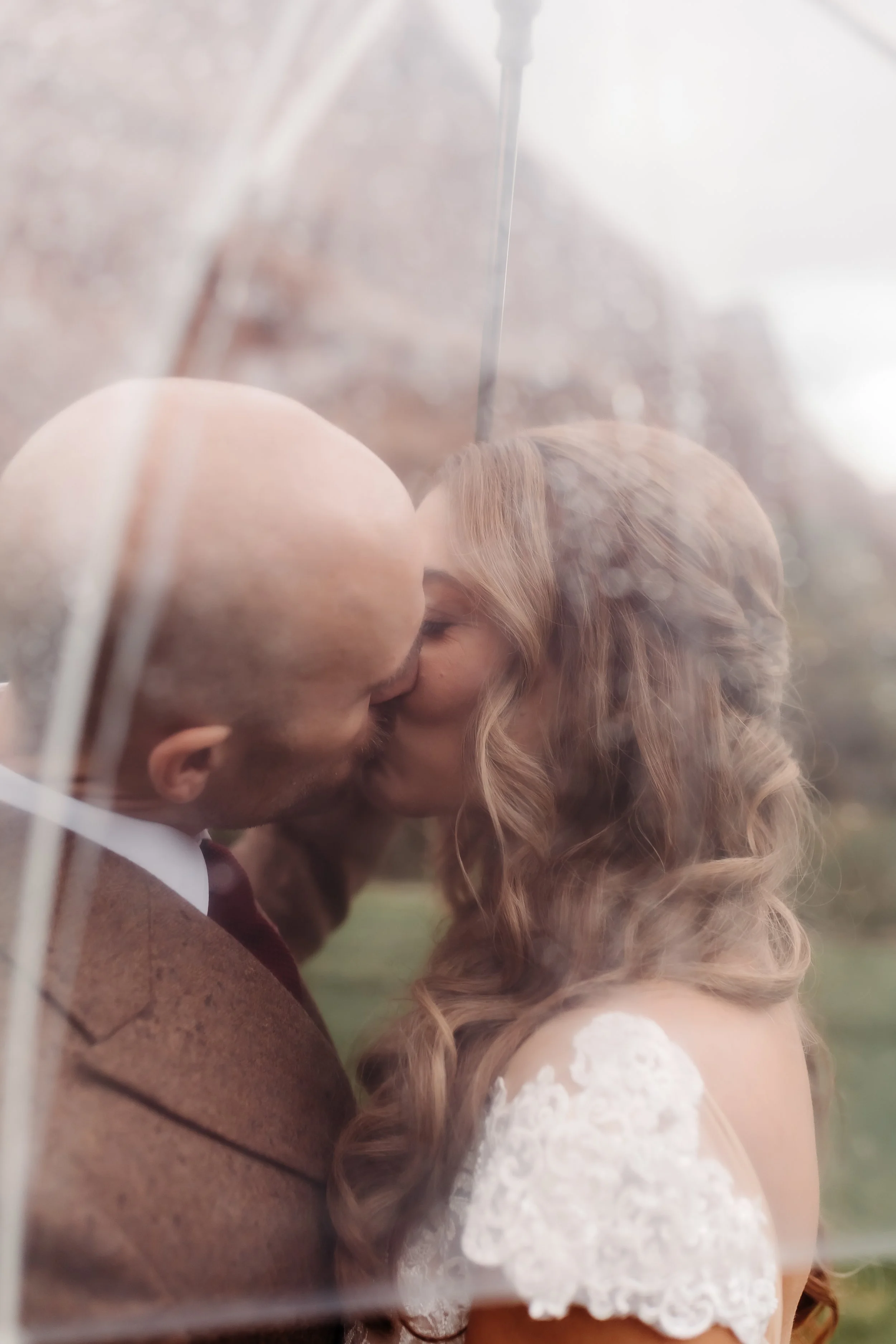 A couple with a clear umbrella sharing a kiss on a rainy day, the woman's curly hair and lace wedding dress are visible.