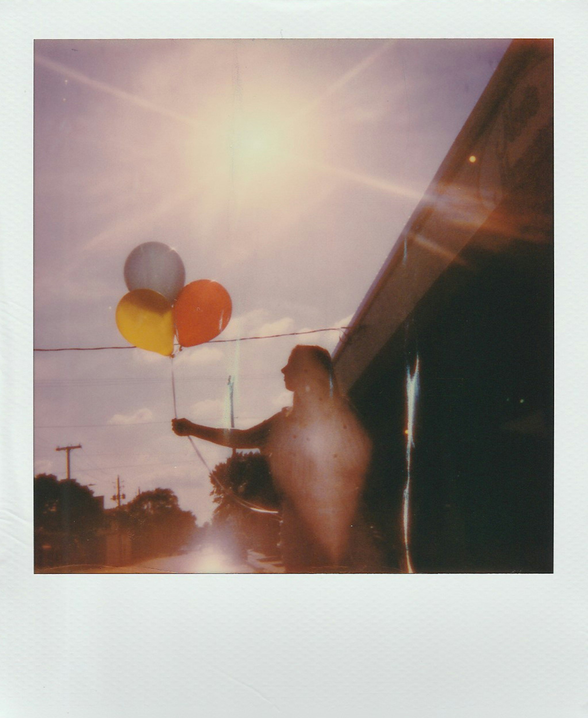A person holding a balloon string with three balloons against a bright sun in the sky, near a building and utility poles.