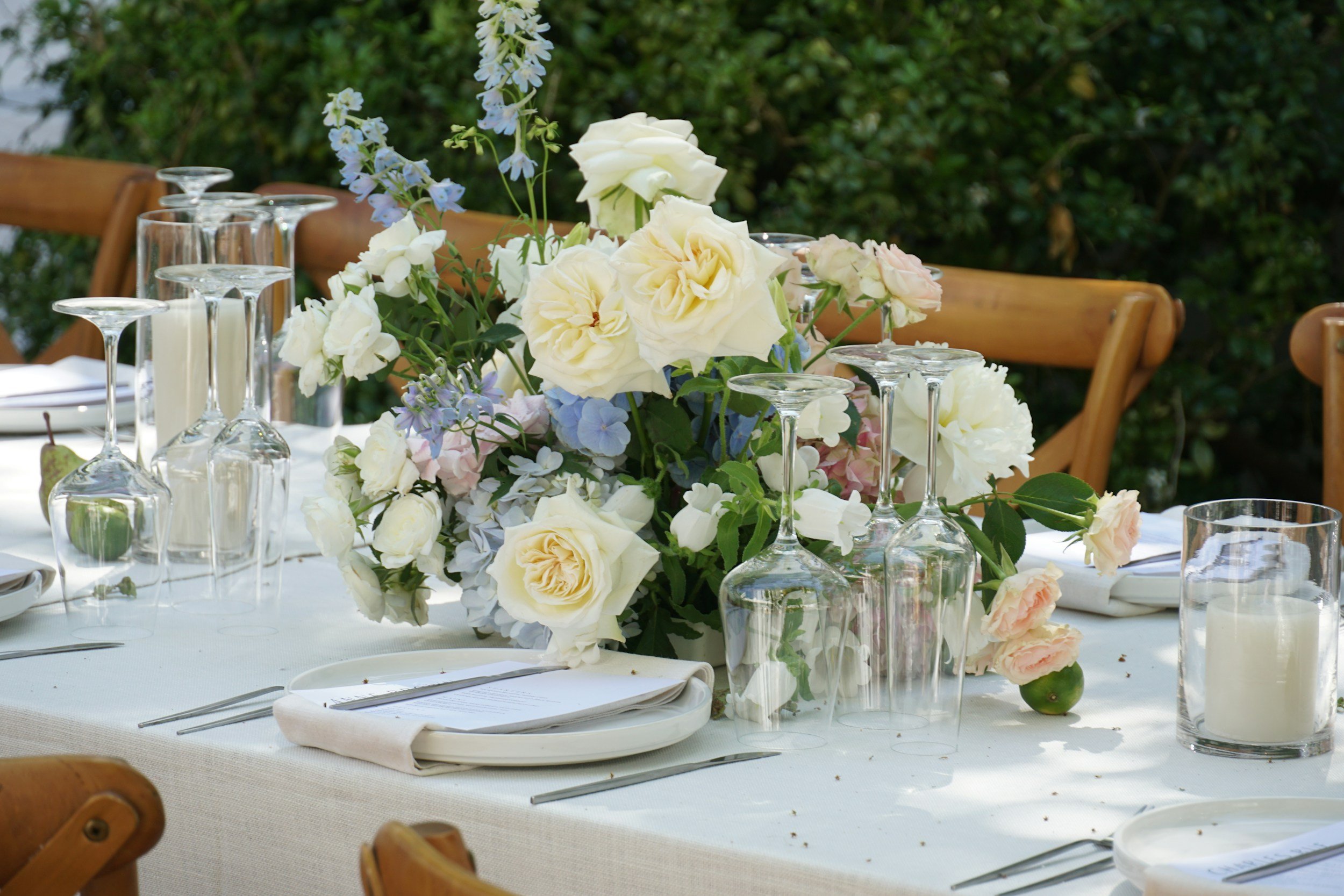 Elegant outdoor dining table with a white tablecloth, floral centerpiece with white and pastel roses, hydrangeas, and greenery, surrounded by empty wine glasses, candles, and place settings, in a garden setting.