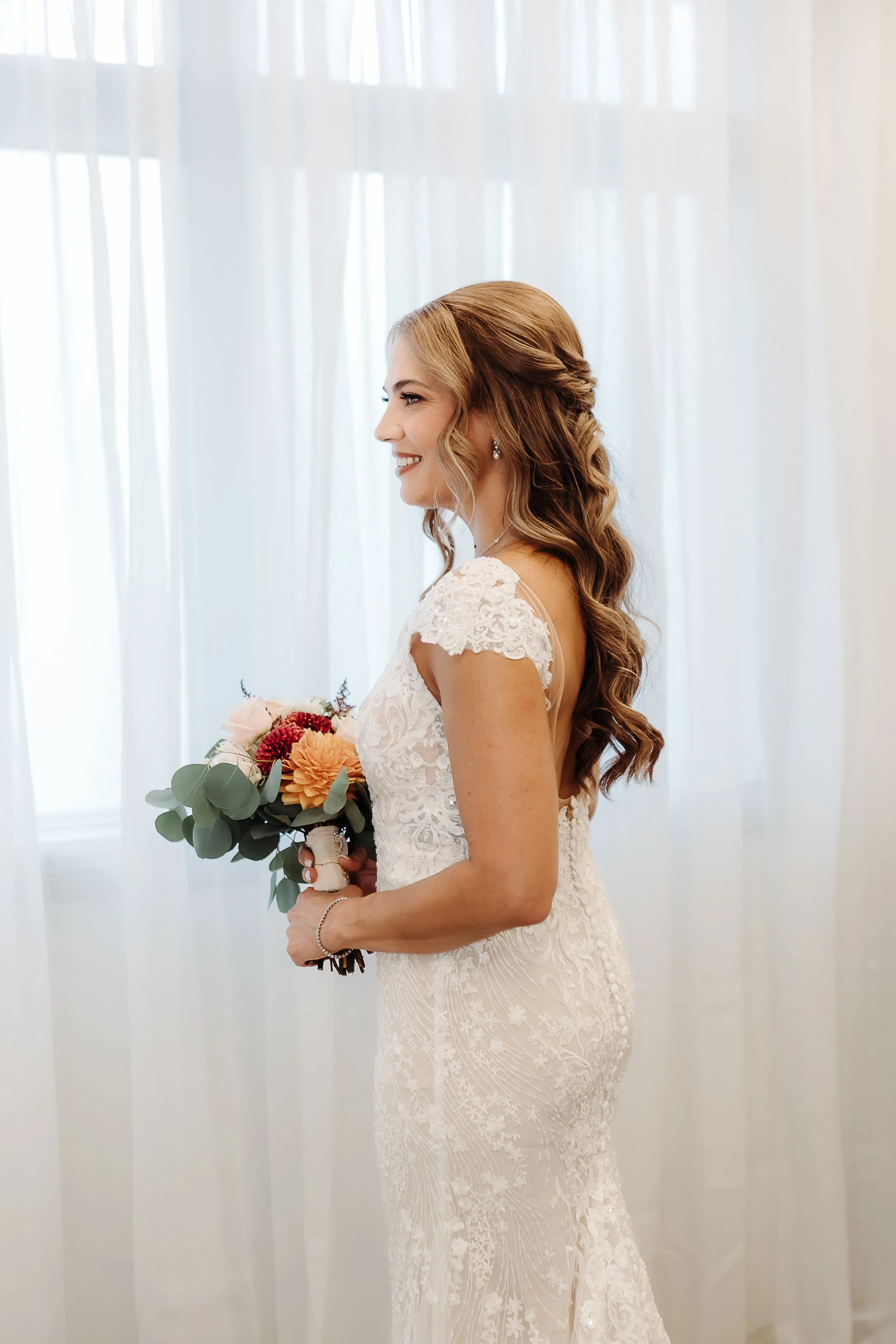 A bride with long, wavy hair in a wedding dress holding a bouquet of pink, red, and orange flowers, standing in front of sheer curtains.