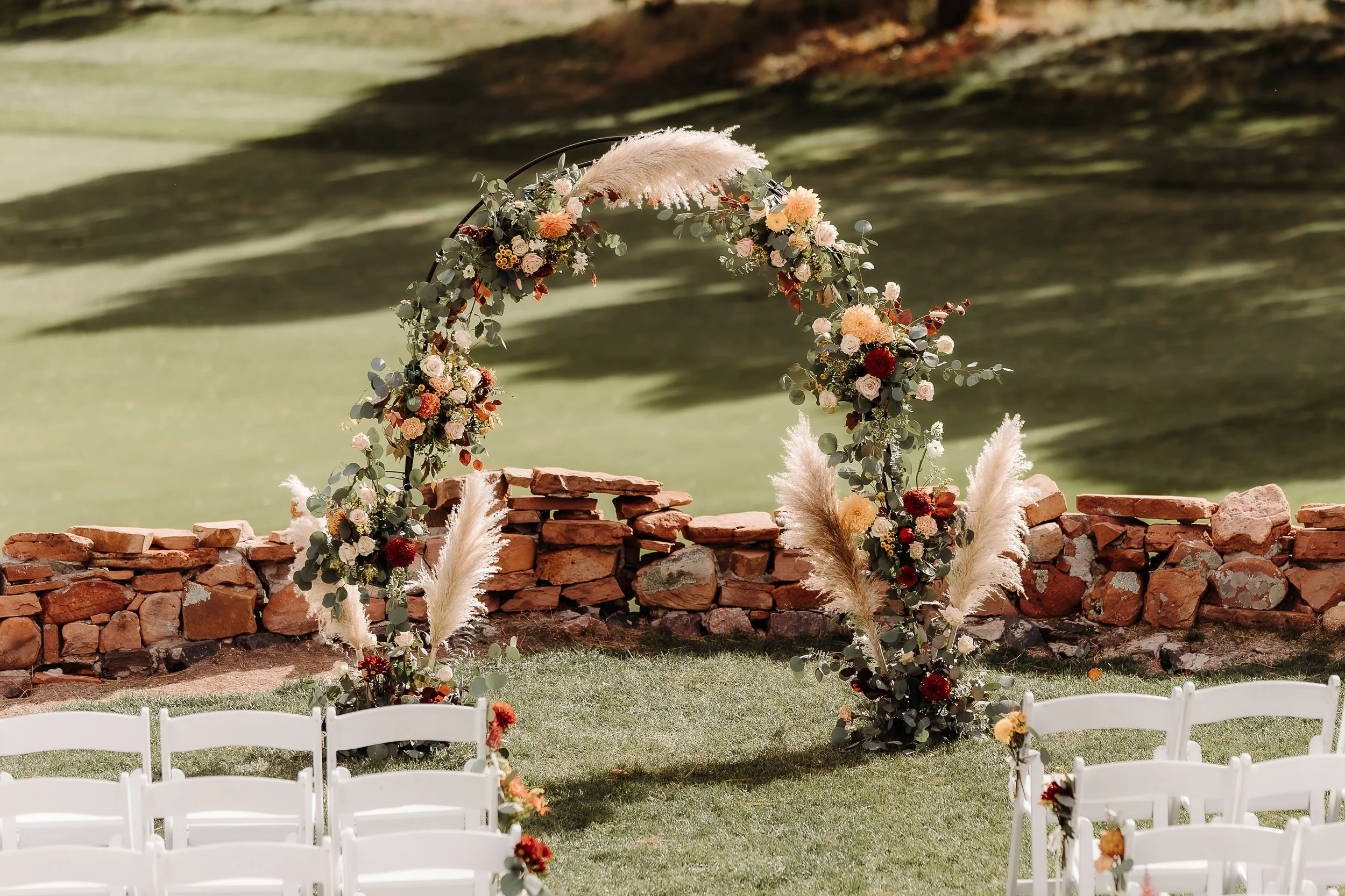 Outdoor wedding ceremony setup with a floral arch decorated with pink, white, and red flowers, and pampas grass, set in front of a stone wall on a grassy area.