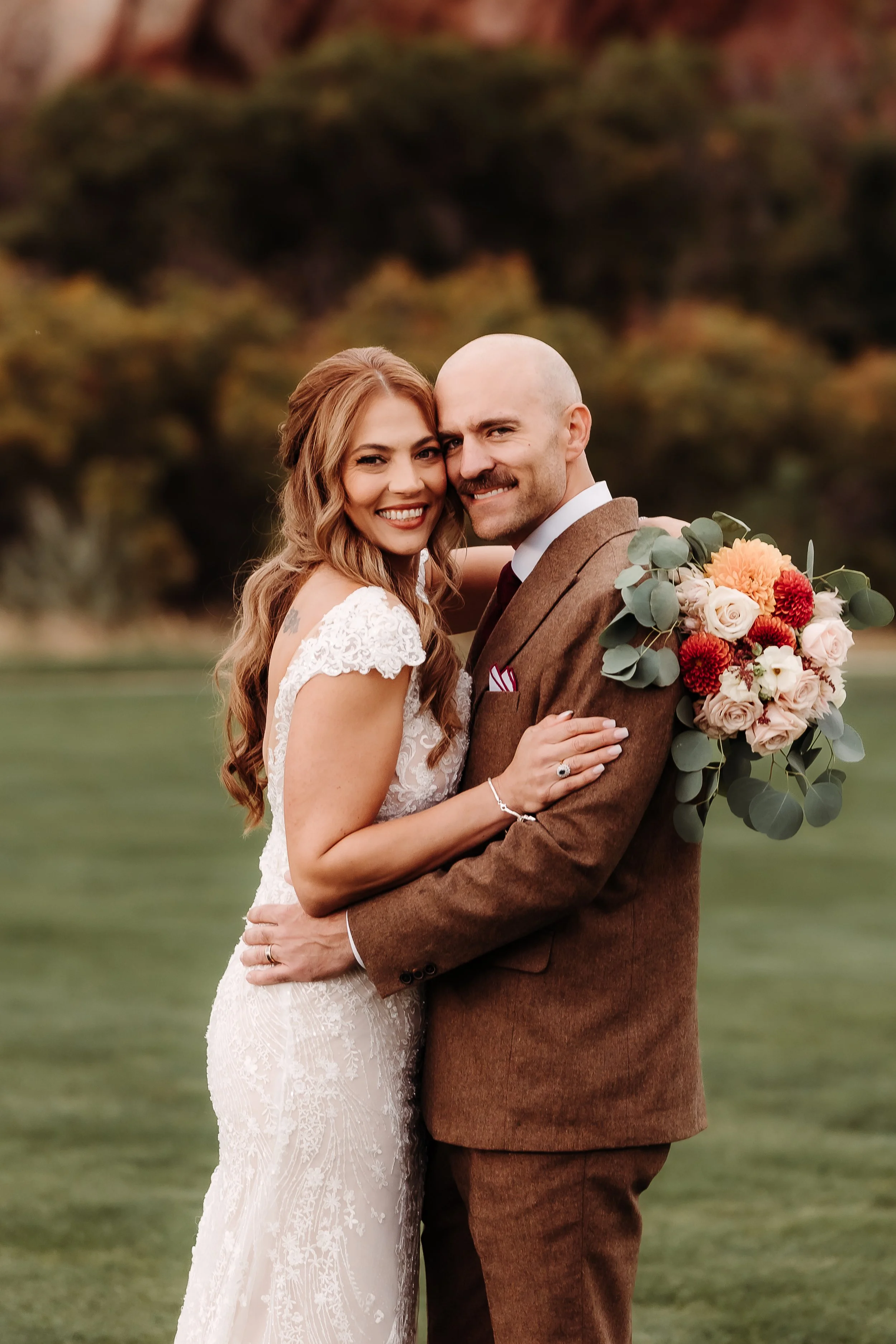 A newlywed couple smiling and embracing outdoors, with the woman wearing a white lace wedding dress and the man in a brown suit, holding a bouquet of flowers, against a blurred, natural background.