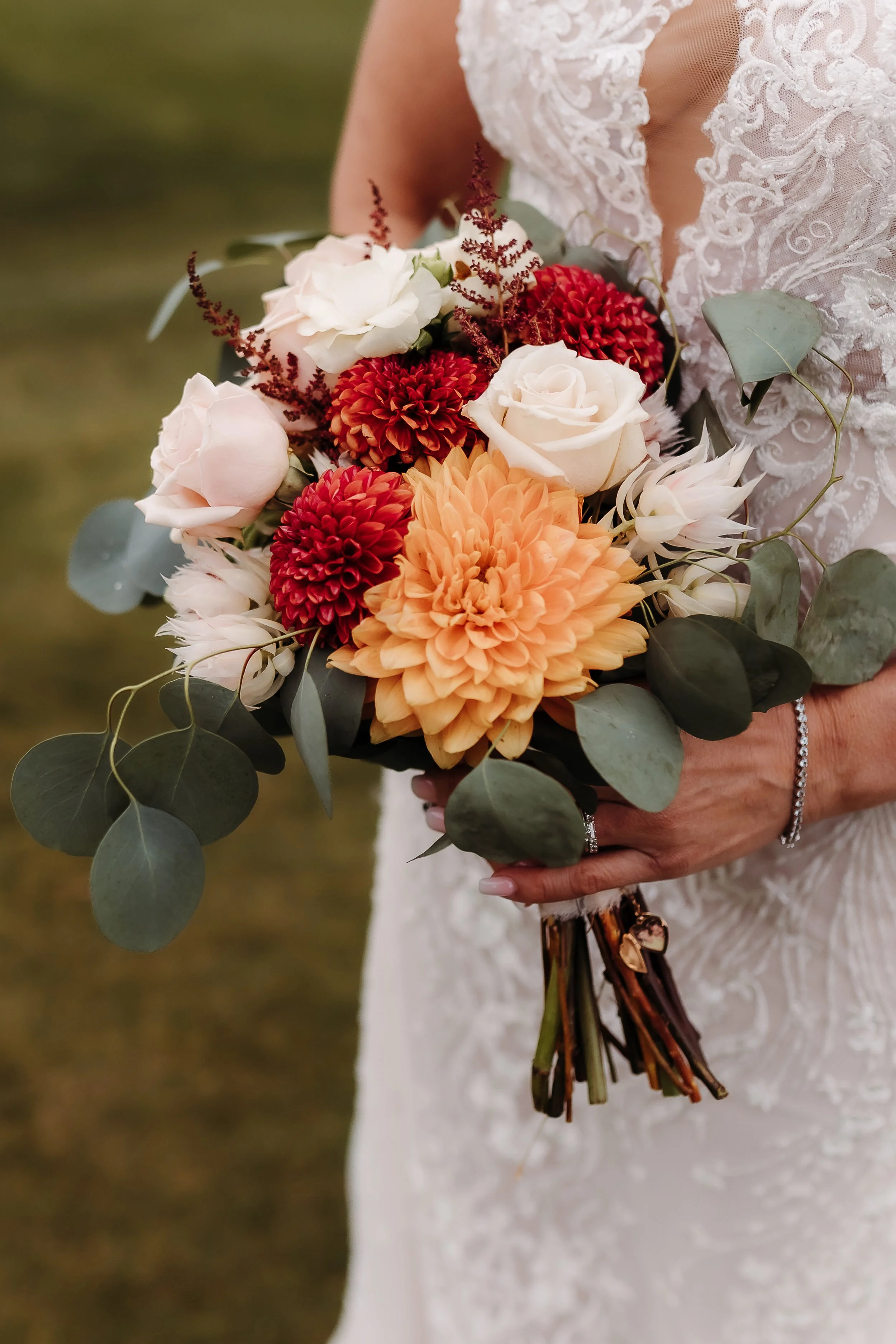 A bride in a lace wedding dress holding a bouquet of peach, white, and red flowers with green leaves.