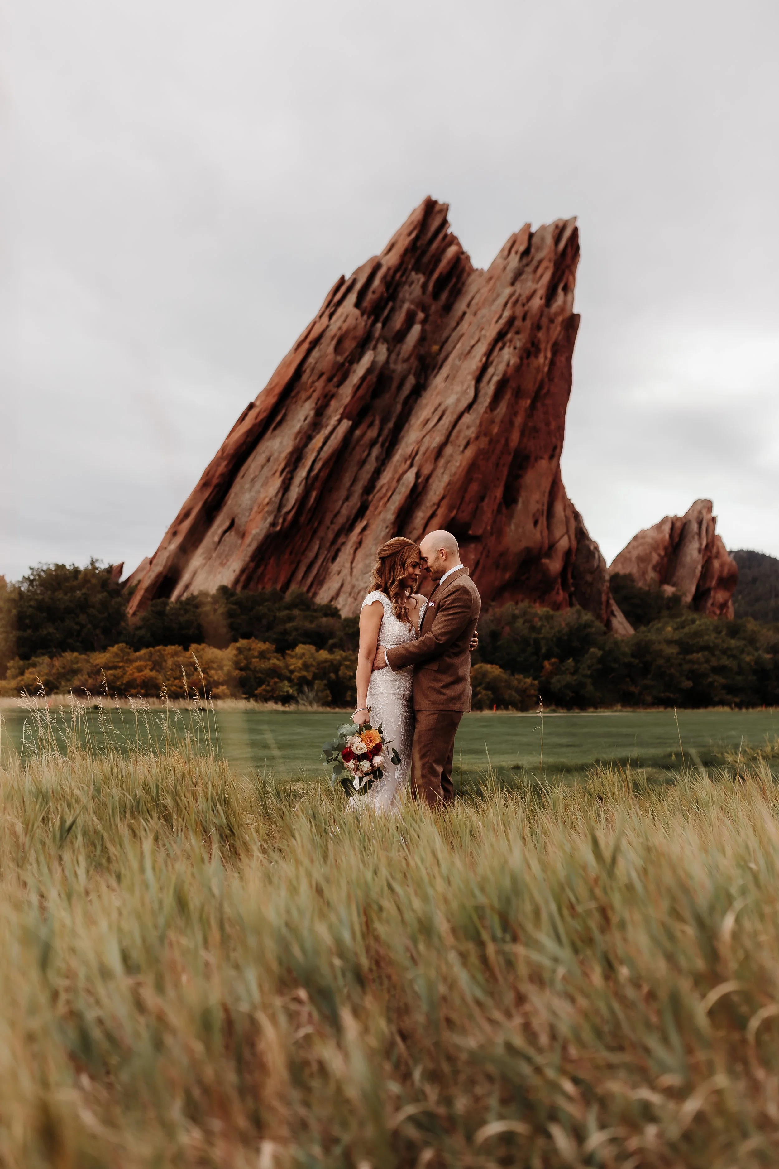 A bride and groom in wedding attire standing close together in a grassy field with a large rock formation and trees in the background.