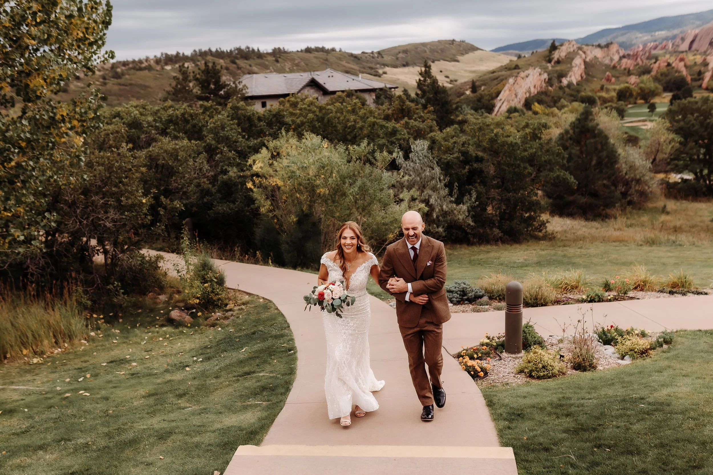 A bride in a white lace wedding dress walking arm-in-arm with a man in a brown suit on a curved sidewalk outdoors. The bride is holding a bouquet of pink and white flowers. They are smiling and laughing as they walk.