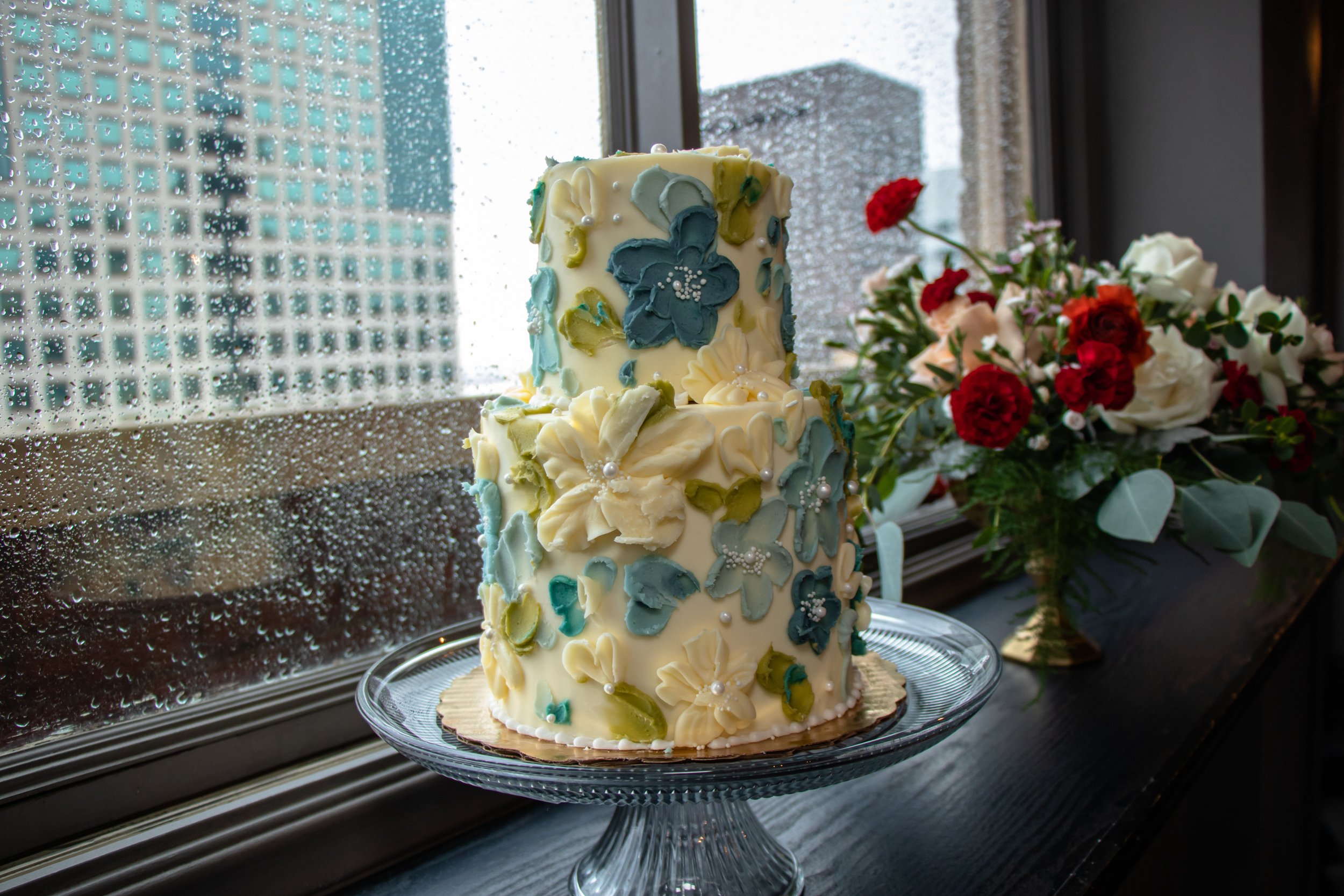 A tall, multi-tiered wedding cake decorated with cream-colored and blue flower patterns, topped with sugar flowers and edible pearls, placed on a glass stand by a window with rain droplets and a cityscape background. A bouquet of red and white flowers is next to the cake.