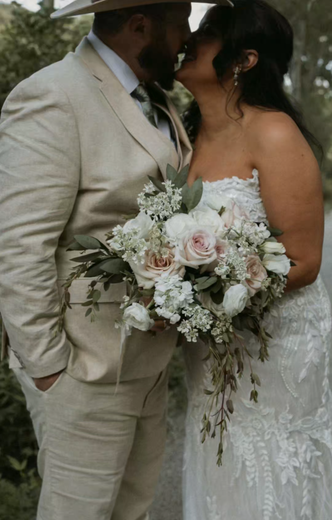 A couple dressed in wedding attire sharing a kiss, with the bride holding a bouquet of white and blush roses and greenery.