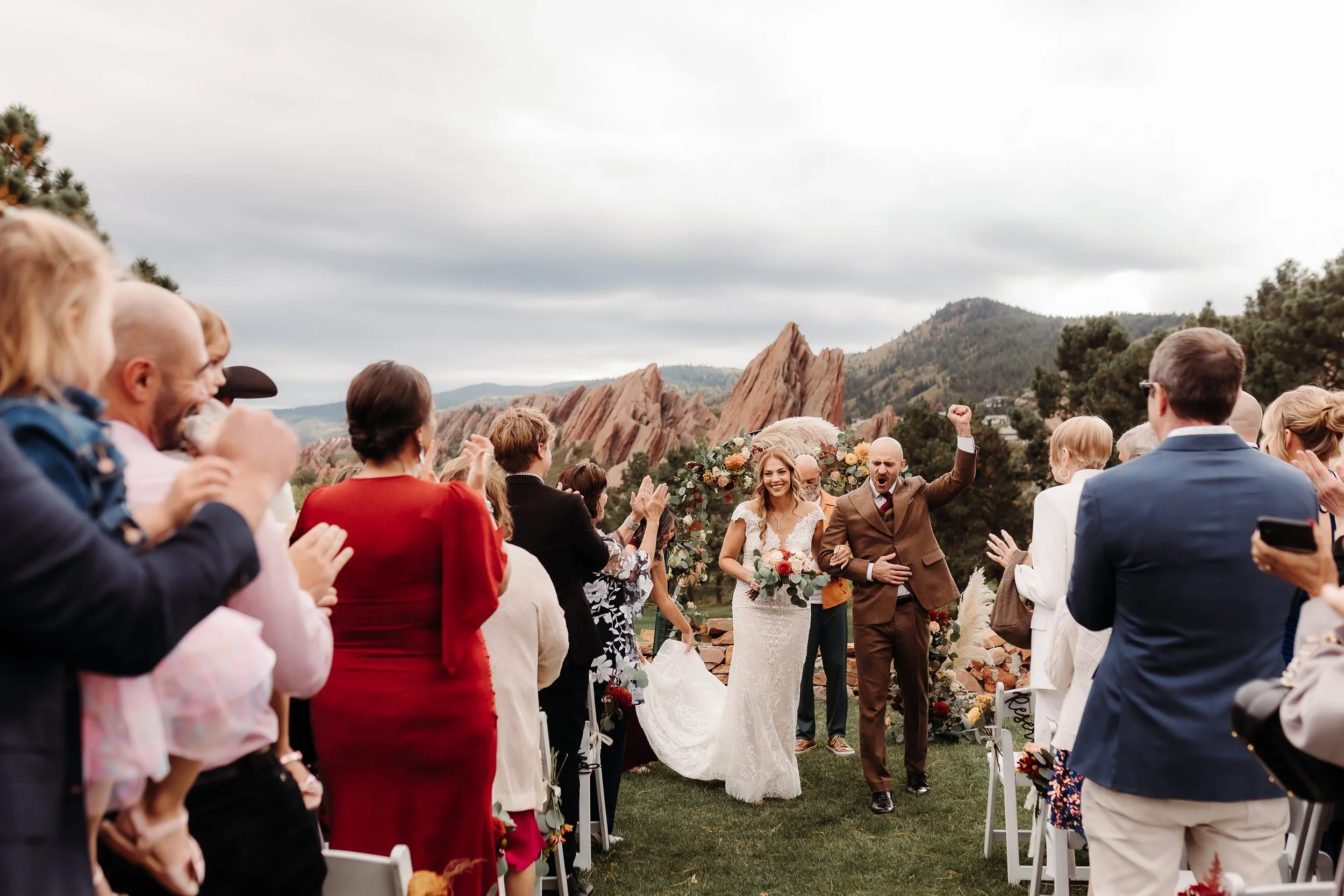 Bride and groom walking down the aisle at an outdoor wedding ceremony, surrounded by smiling guests, with red rock formations and mountains in the background under a cloudy sky.