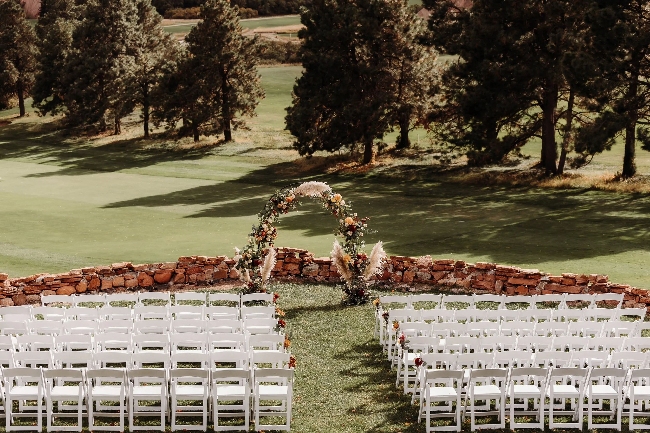 Outdoor wedding setup with white chairs arranged in rows facing a floral arch, on a grassy area near a stone wall, with trees and a field in the background.