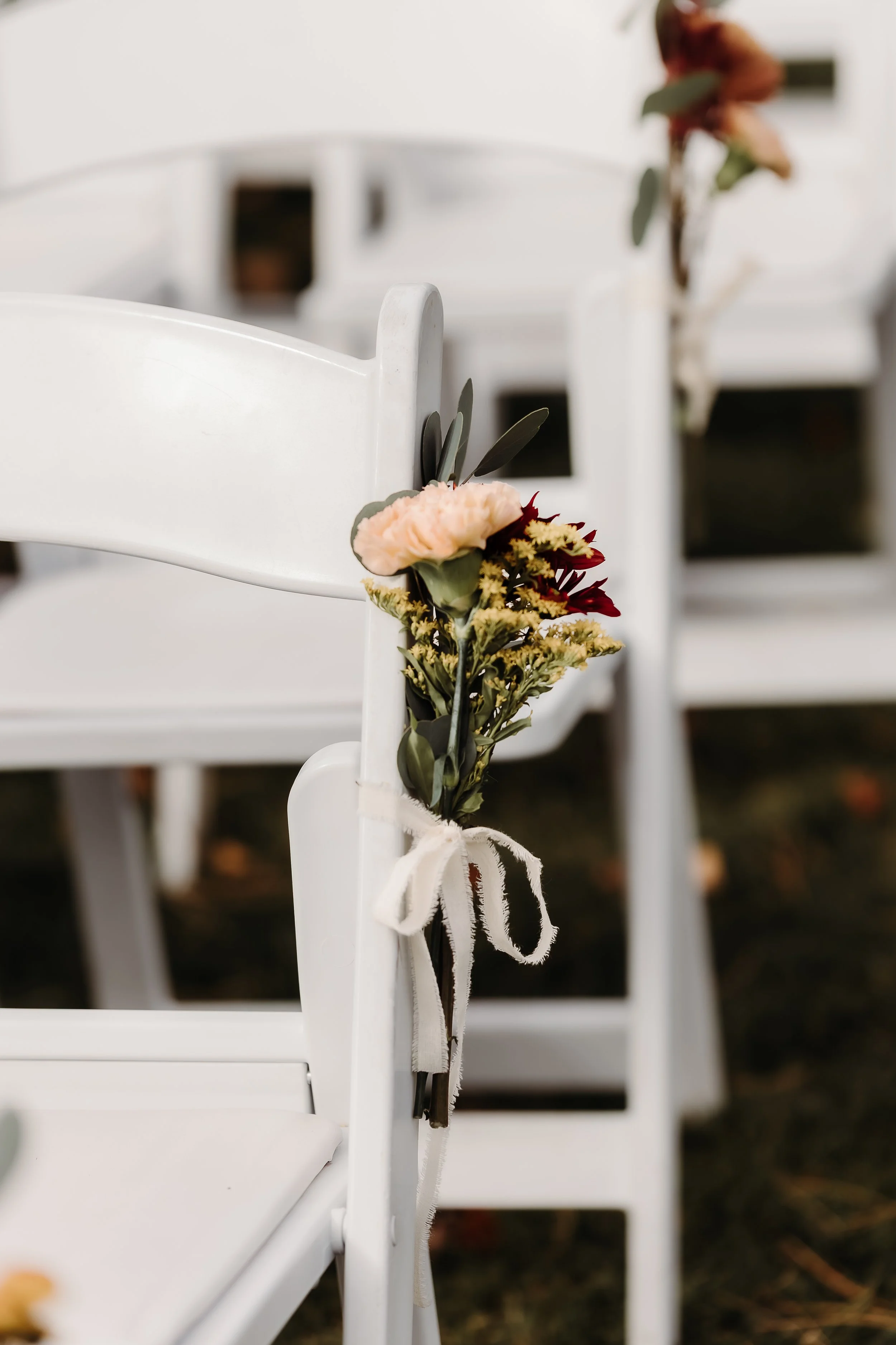 Close-up of a white wedding chair decorated with a small floral arrangement tied with a white ribbon, set outdoors.