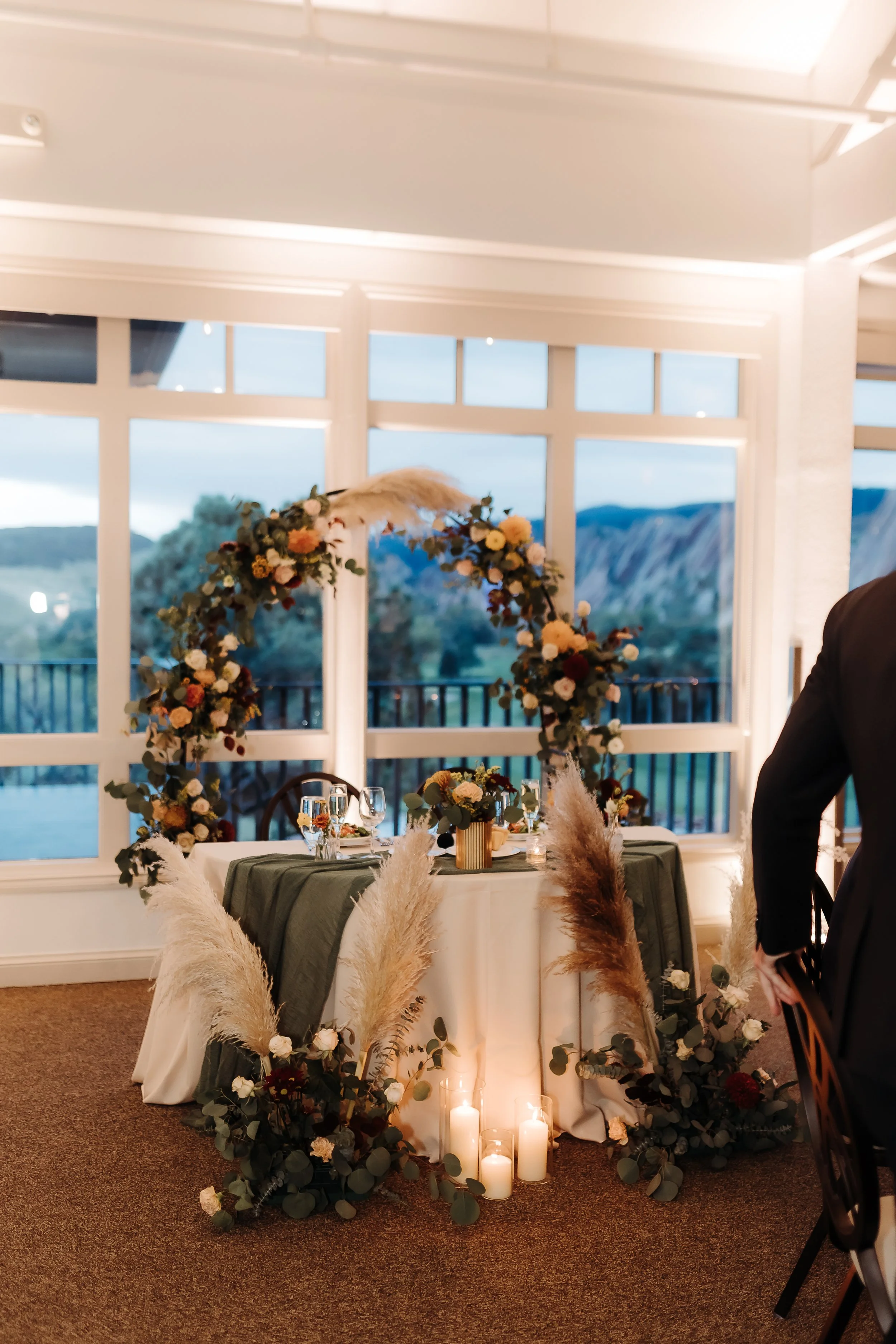 Decorated wedding reception table with candles, floral arrangements, and a large floral arch behind it, set in a bright room with large windows showing an outdoor landscape.