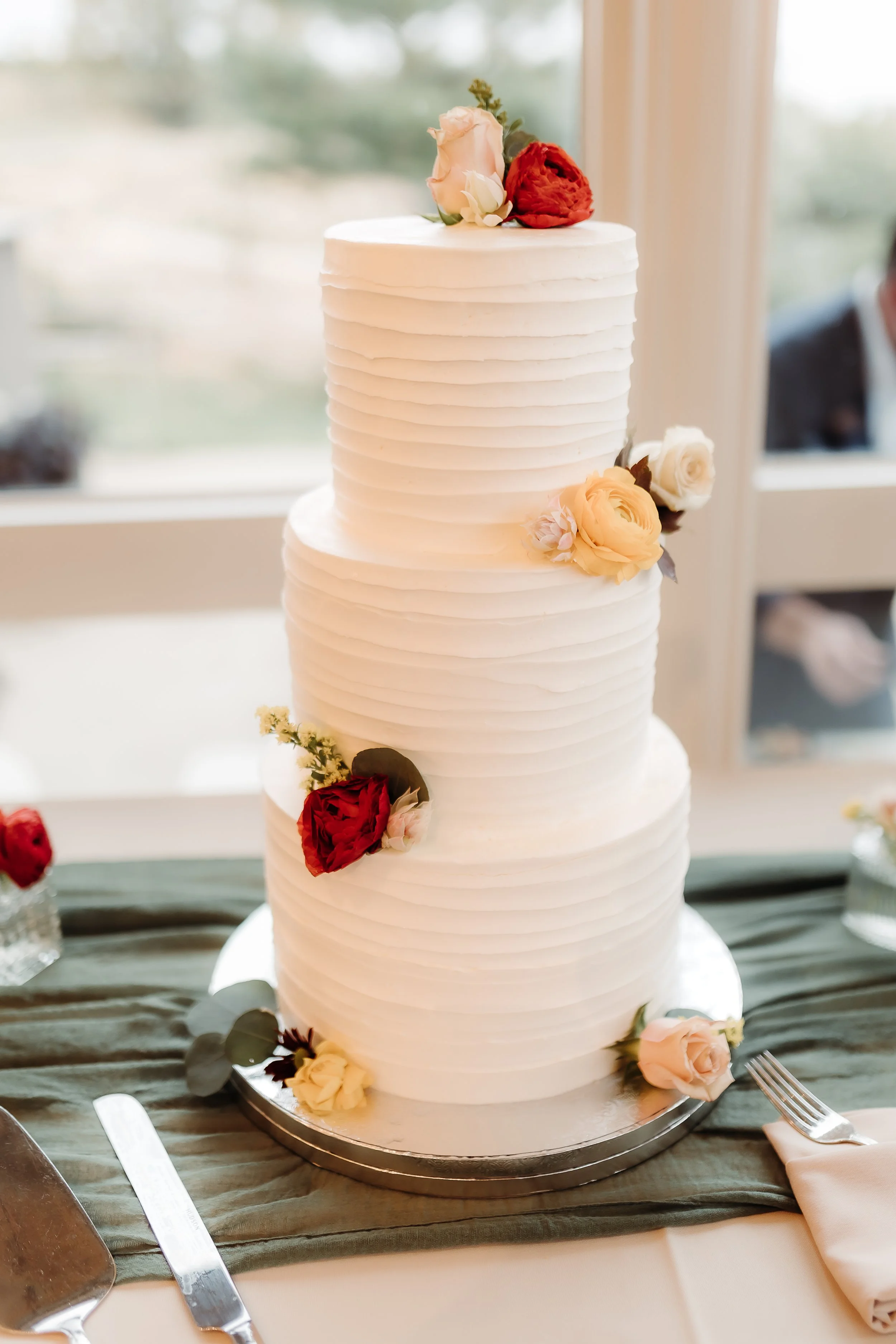 Three-tier white wedding cake with textured horizontal lines, decorated with assorted flowers including roses in shades of white, peach, and red, placed on a silver cake board on a green cloth table runner.