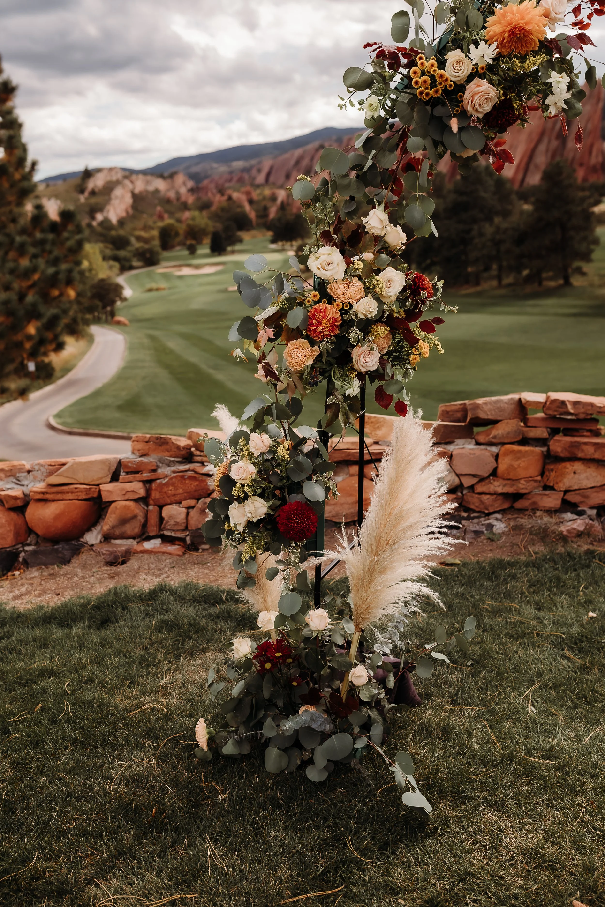 A floral arrangement with white, red, and peach flowers, including roses and dahlias, and pampas grass, set outdoors on a grassy area with a stone wall and a golf course in the background, under a cloudy sky.