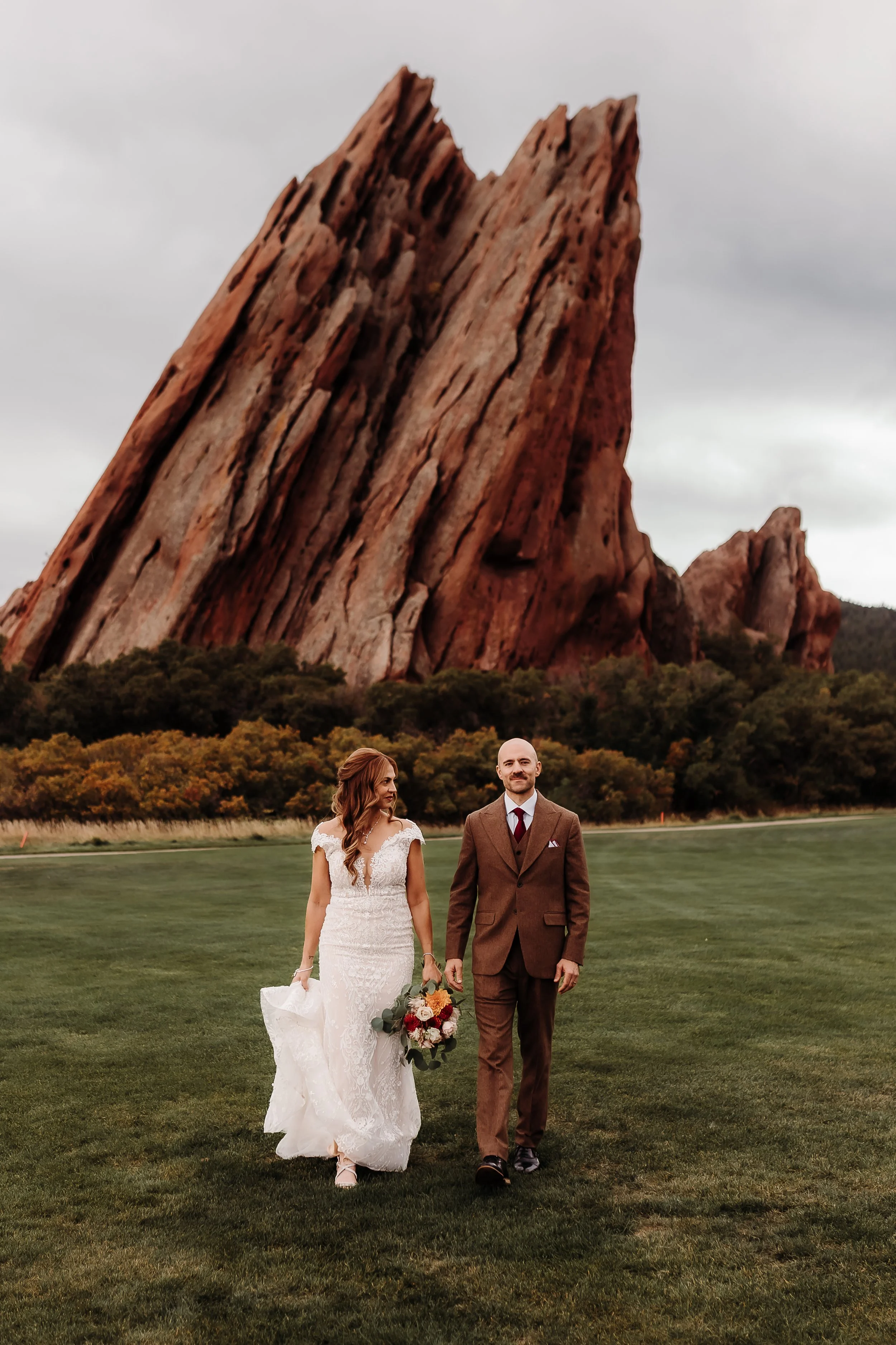 A bride and groom walking on a grassy field with a large, rocky mountain formation in the background.