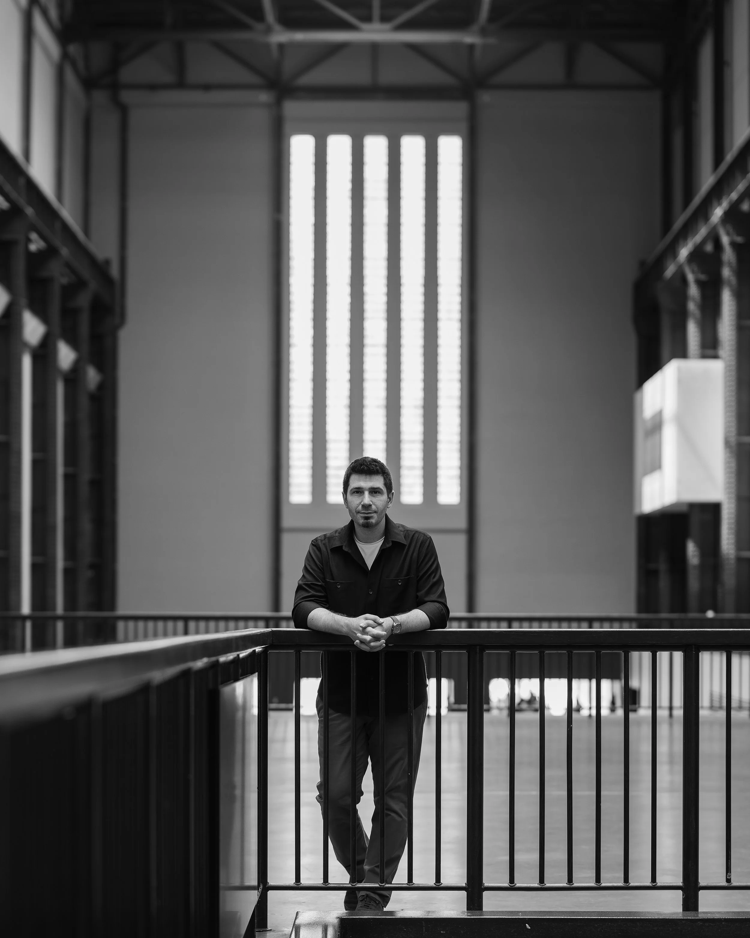 A man, our founder, standing with arms crossed behind a black iron railing inside a large building with high ceilings and a tall window with vertical panels in the background.