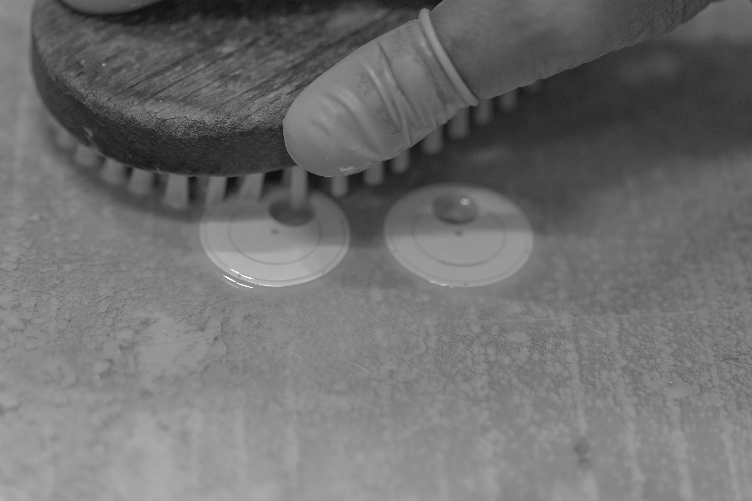 Close-up of a hand with rubber glove on, holding a wire brush, applying grenage onto the N01 dials. The image is in black and white.