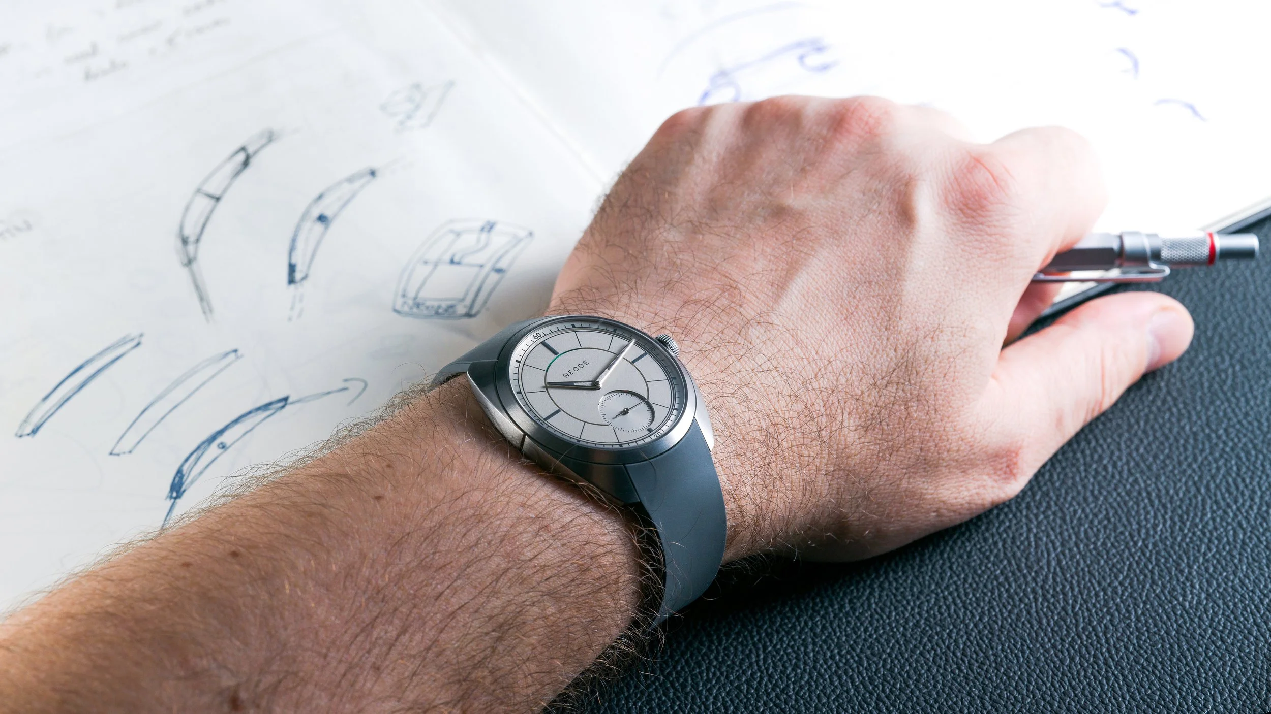 A person's arm wearing a titanium N01 timepiece with a light gray face, resting on a blue textured surface, with sketches of watch profiles and designs on white paper in the background.