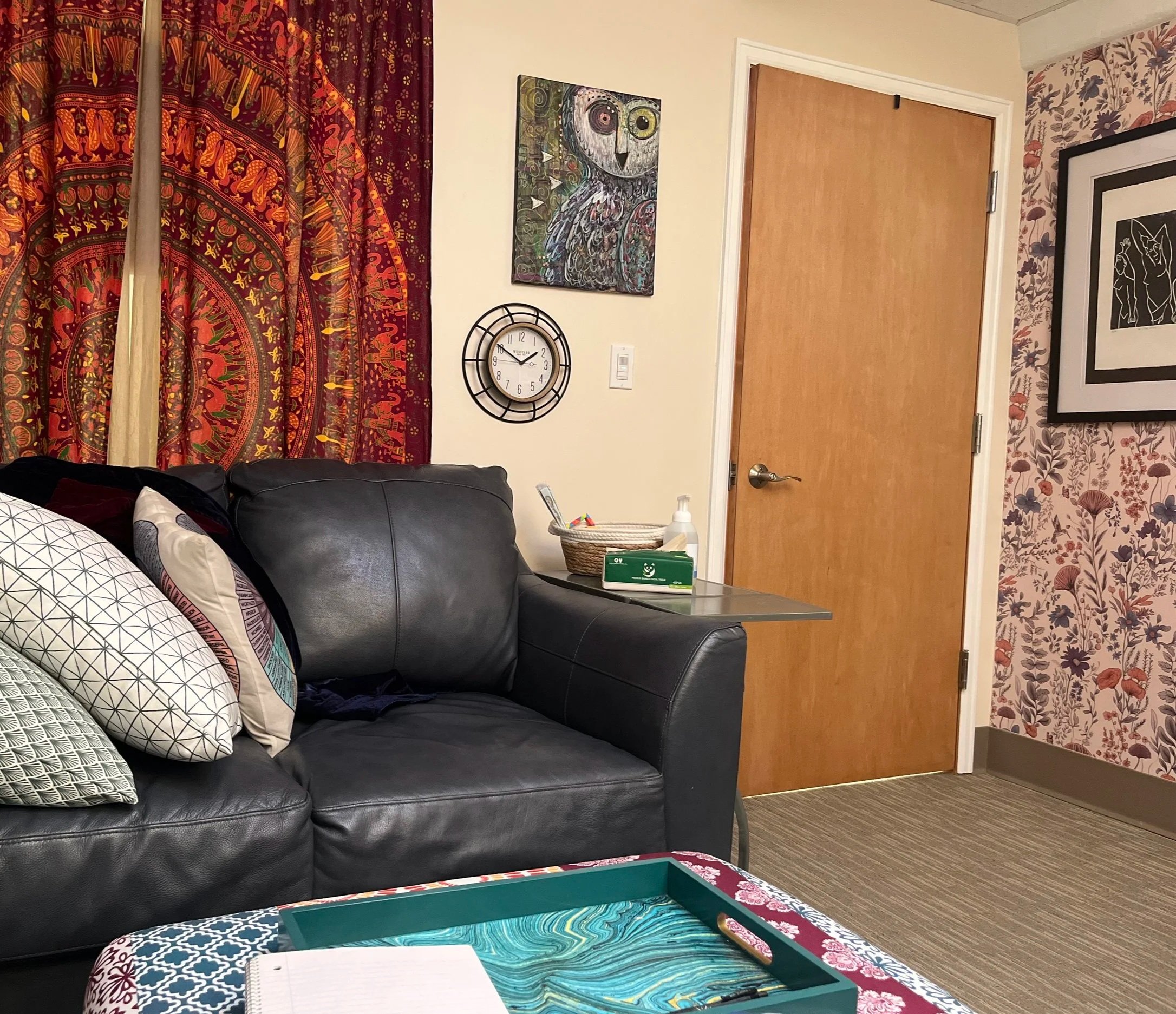 A cozy room corner featuring a black leather sofa with patterned pillows, a small side table with fidget toys, tissue, hand sanitizer, and wall decorations including paintings and a clock. There's a wooden door and floral wallpaper.
