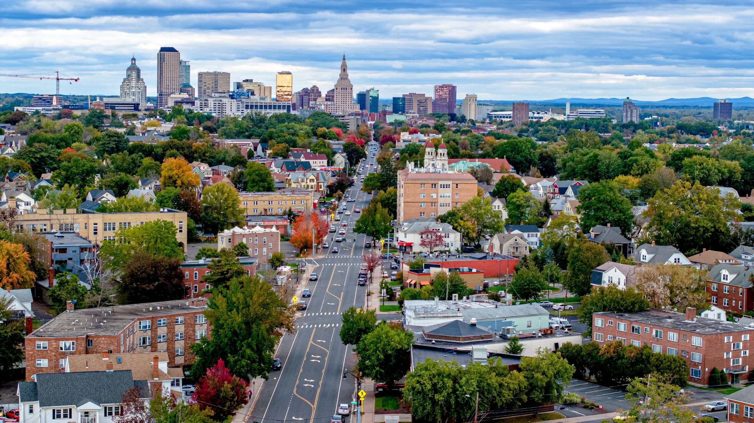 A cityscape with a mix of low-rise and high-rise buildings, tree-lined streets, and a cloudy sky in the background.