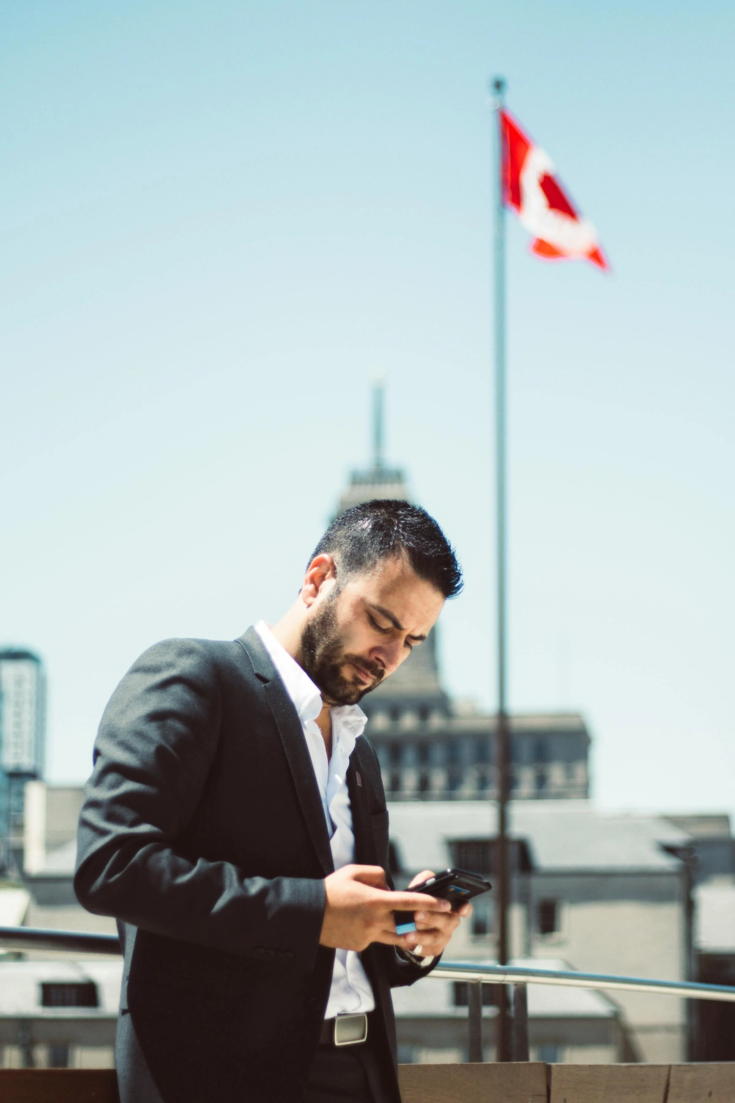 A man in a black suit and white shirt, with a beard and short dark hair, looking down at his phone on a city rooftop. A Canadian flag and a city skyline, including a historic building with a clock tower, are visible in the background.