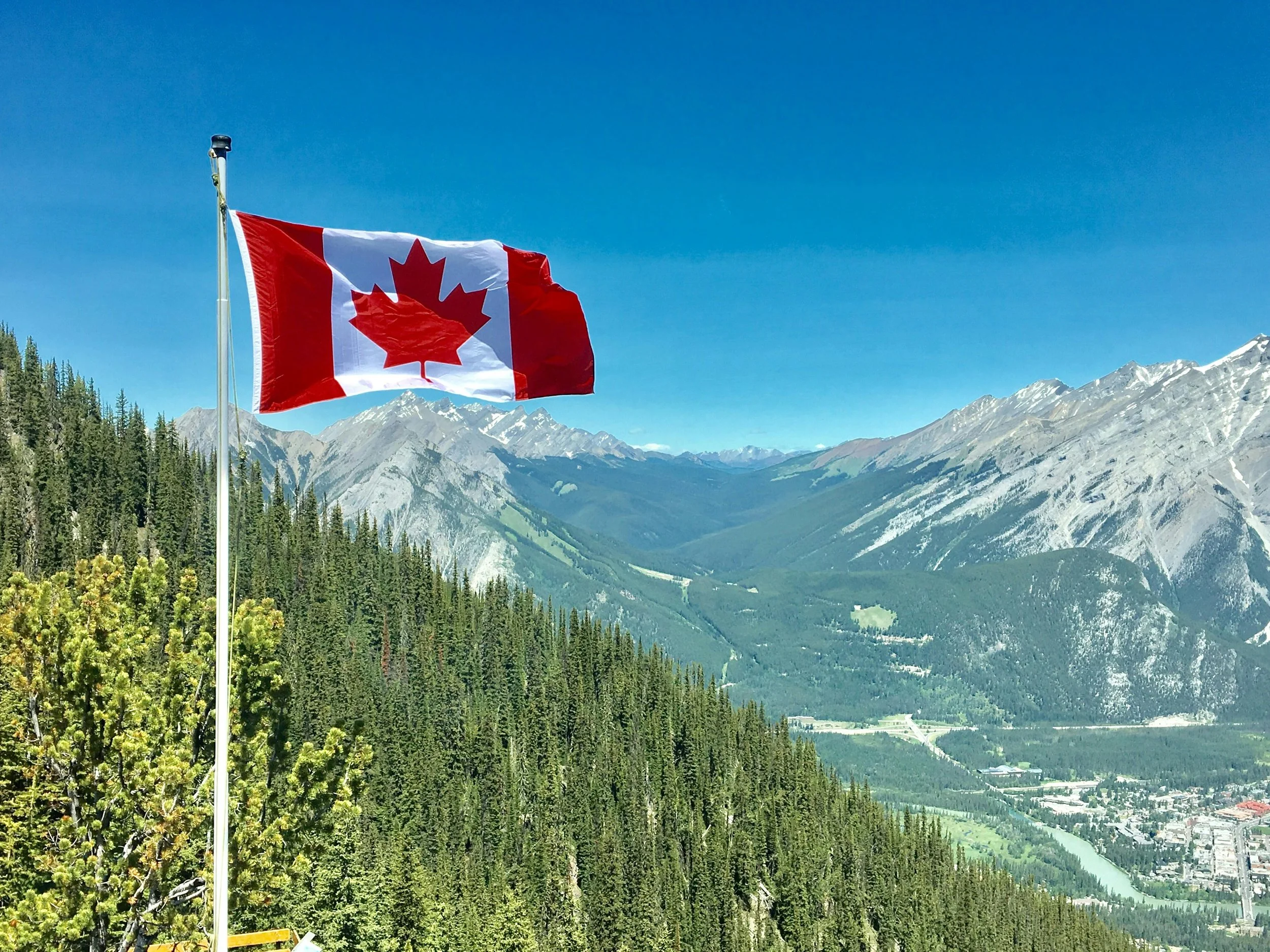 Canadian flag flying on a mountain overlooking a forest, river, and snow-capped mountains under a clear blue sky.