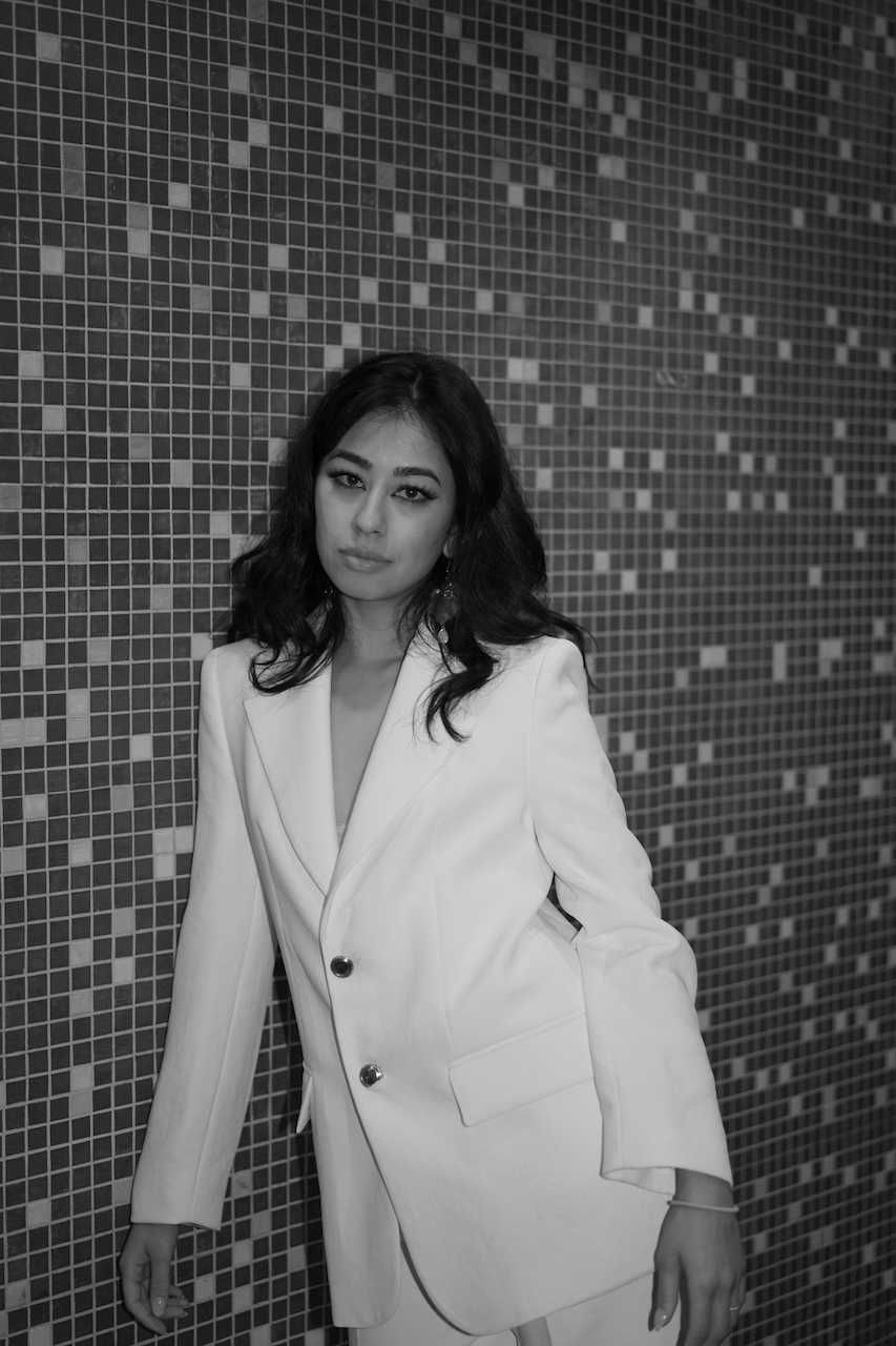 A woman with dark hair and makeup wearing a white blazer standing against a mosaic tiled wall.