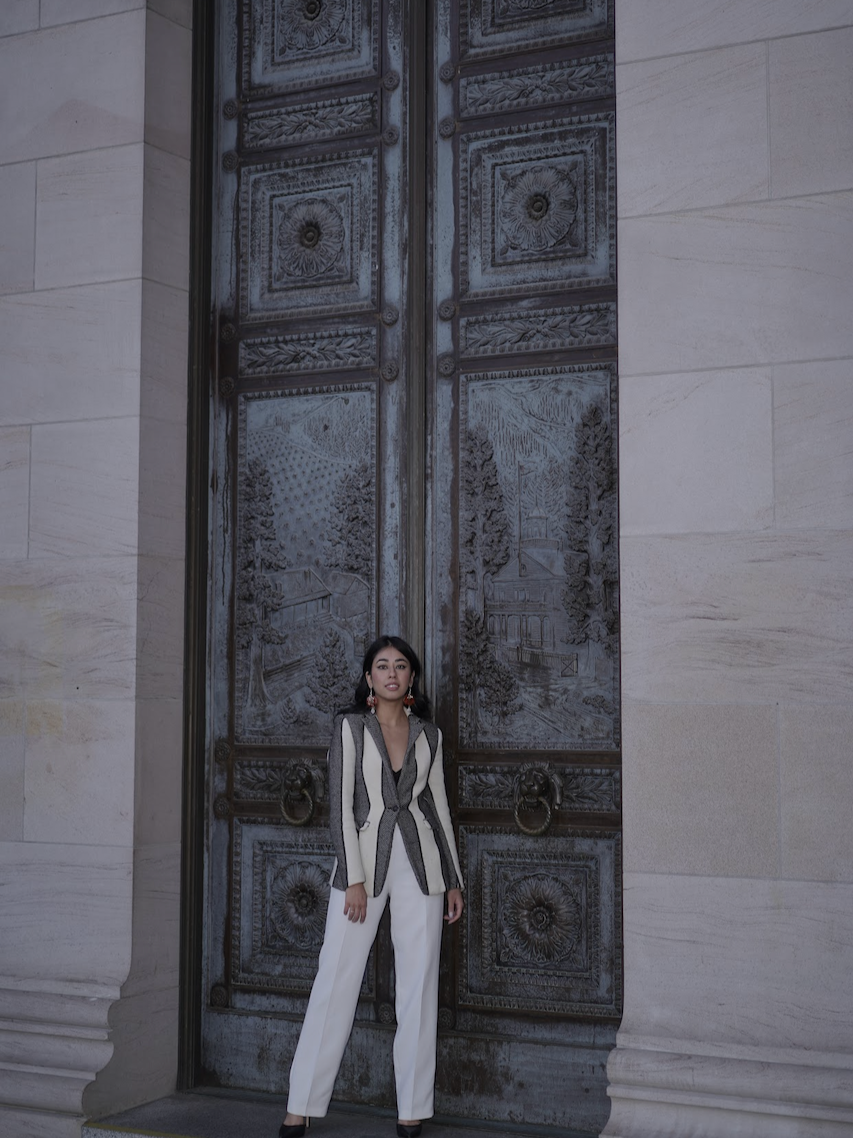 A woman in a stylish black, white, and gray blazer and white pants stands in front of tall, ornate metal doors with detailed engravings.
