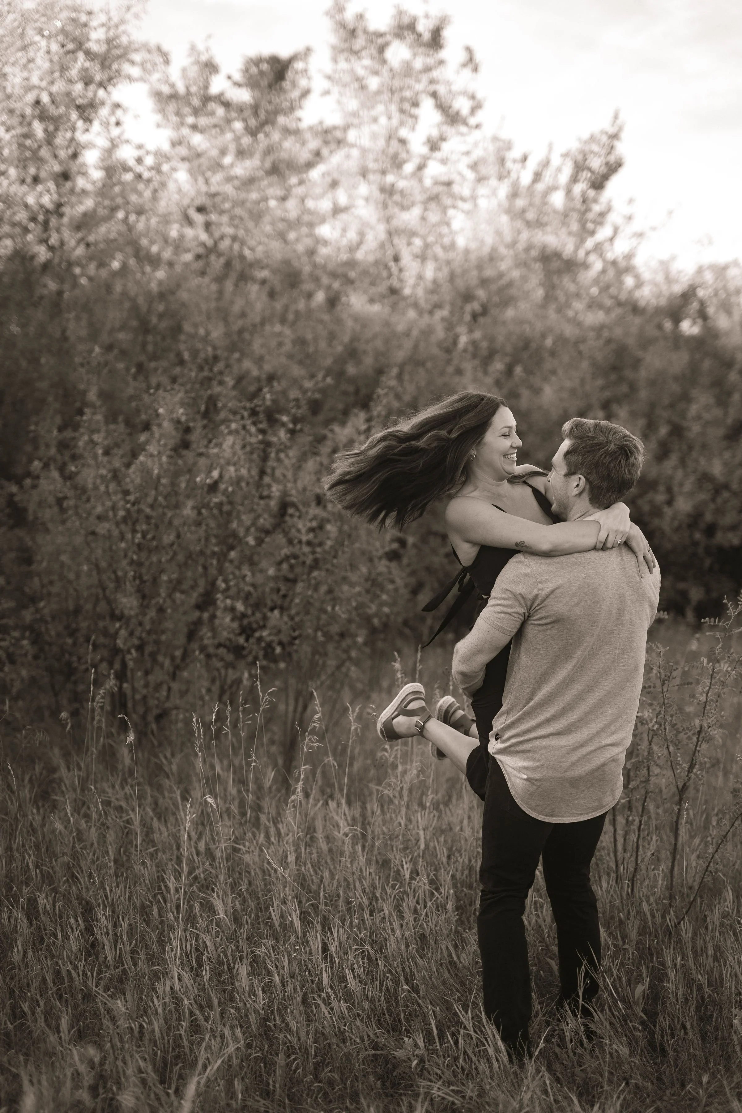 A man lifting a woman in a field with trees in the background, both smiling and appearing happy.