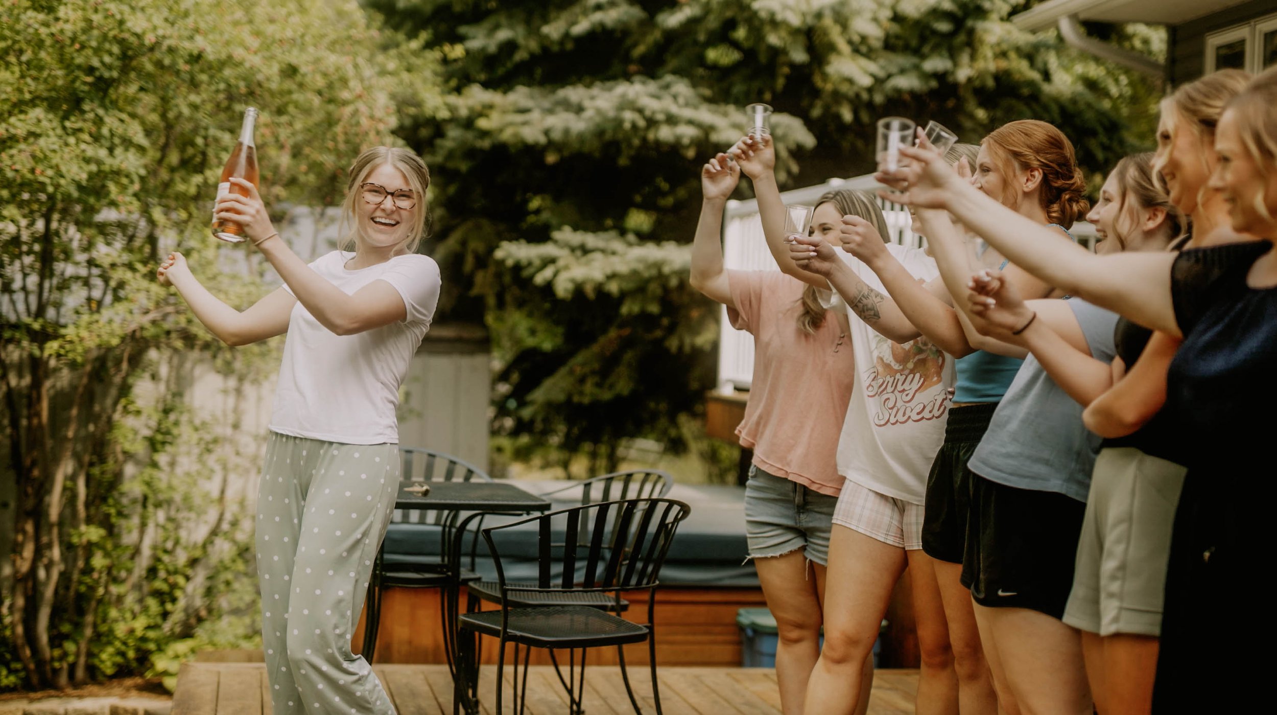 Group of women celebrating outdoors on a wooden deck, holding drinks, with one woman leading a toast. Lush green trees and a hot tub are in the background.