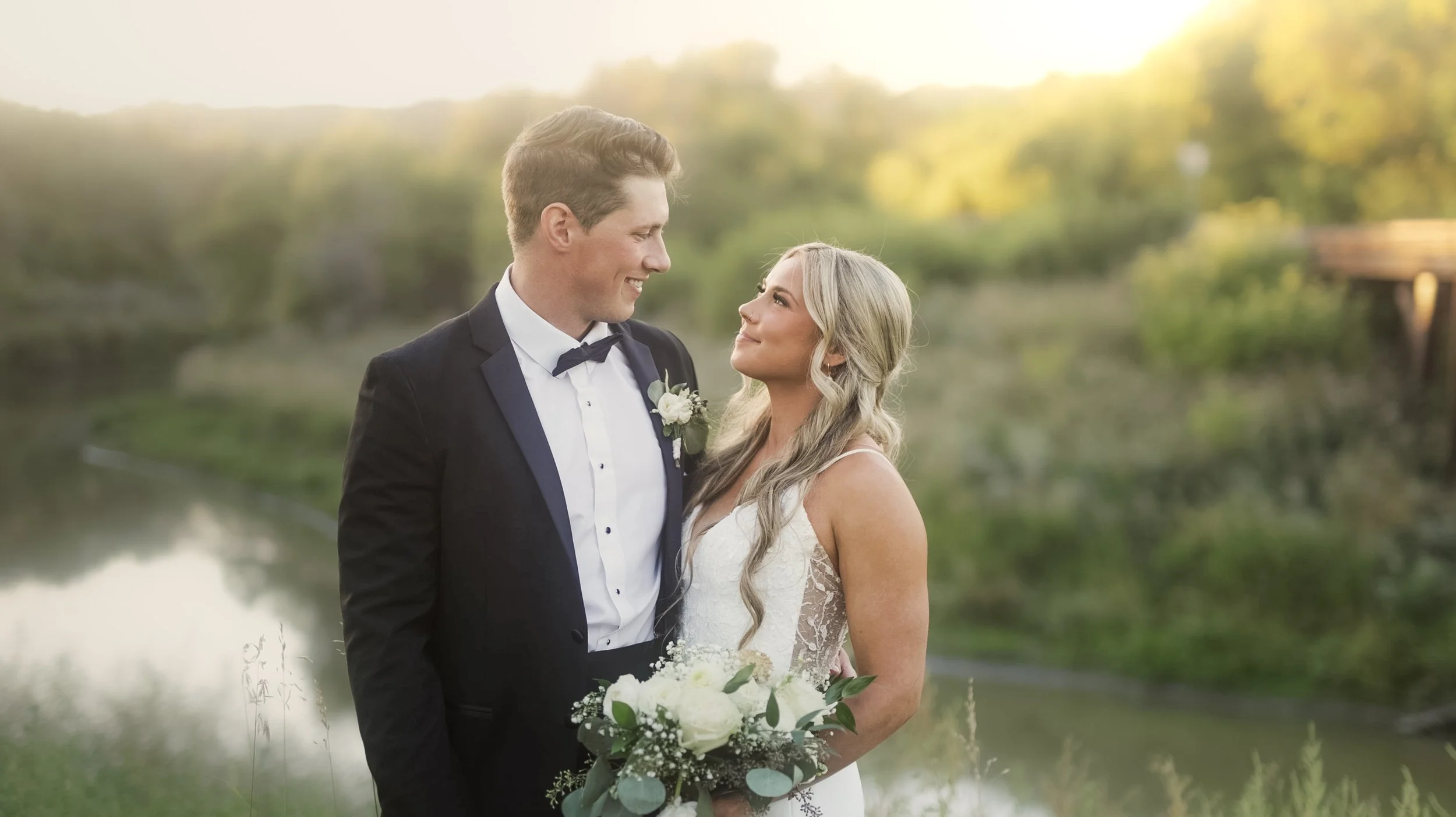 A newlywed couple standing outdoors by a river at sunset, gazing into each other's eyes. The groom is wearing a black tuxedo with a white shirt and bow tie. The bride is holding a bouquet of white flowers and has long, wavy blonde hair. She is wearing a sleeveless lace wedding dress.