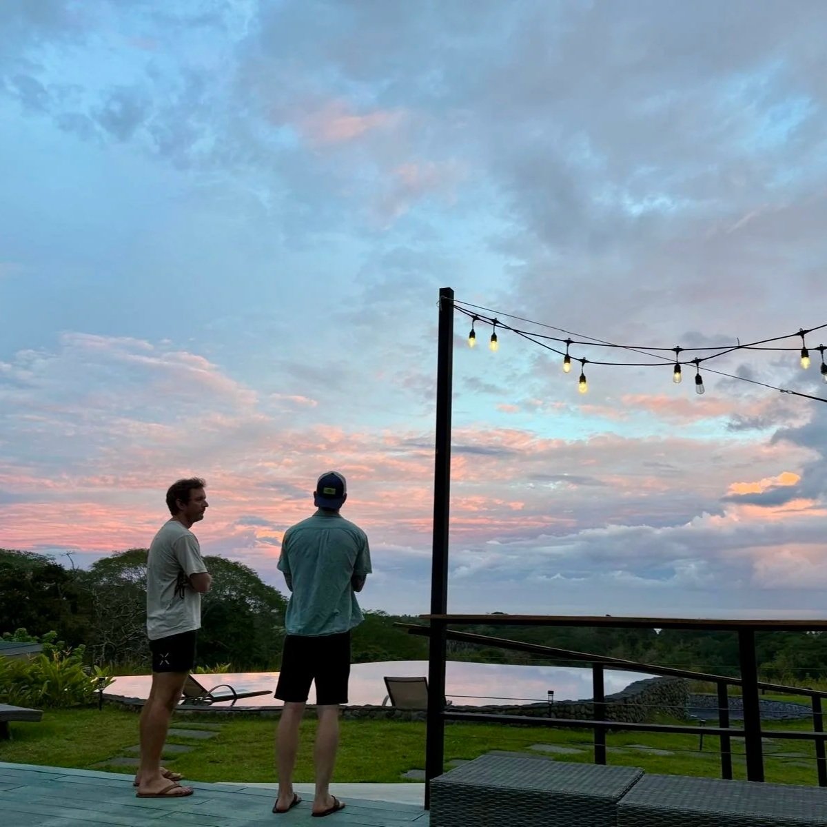 Two men standing and talking outdoors at sunset under a cloudy sky, with string lights hanging overhead, near a railing and a pool.