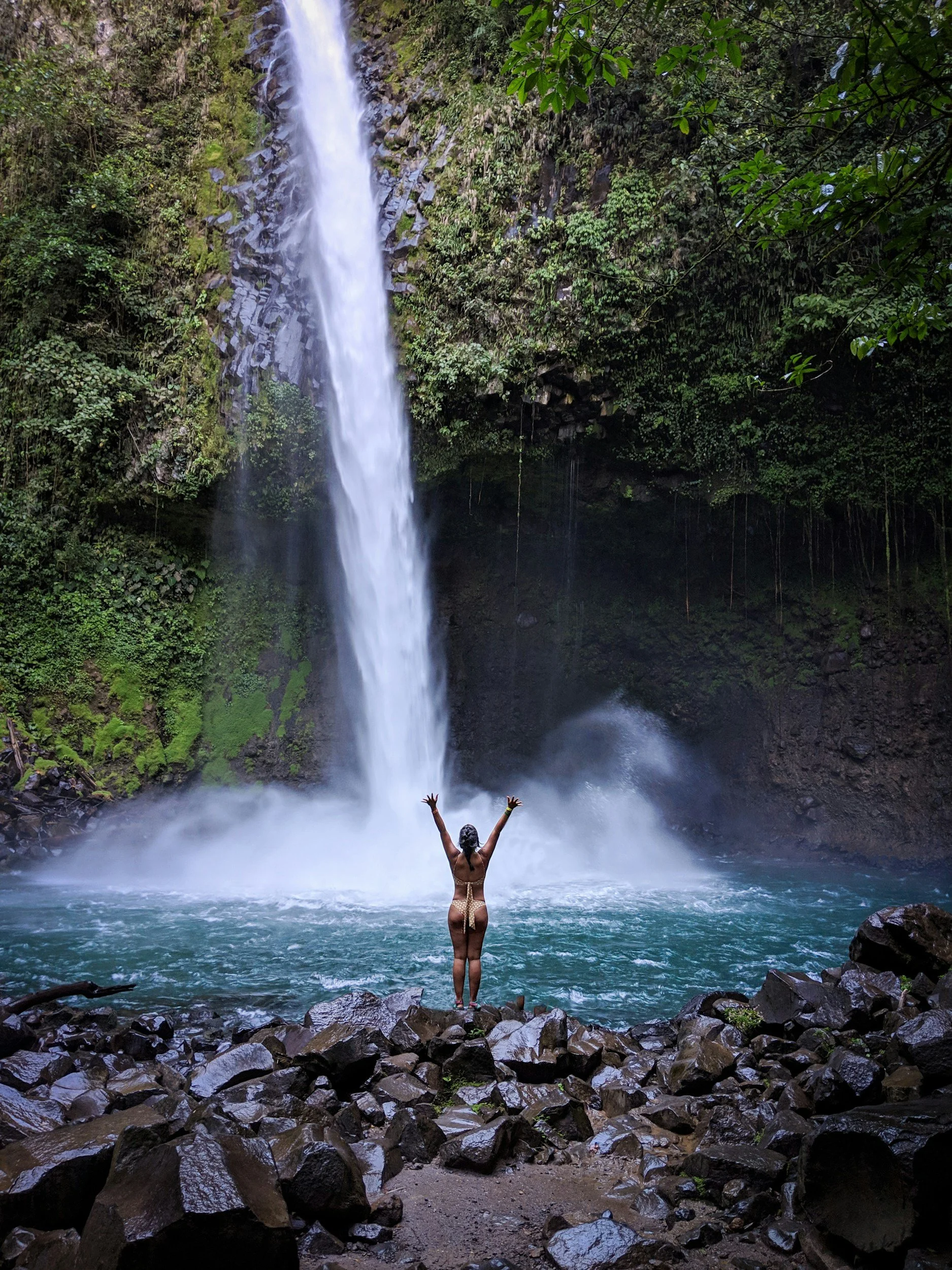 Person standing on rocky ground with arms raised in front of a tall waterfall in a lush, green forest.