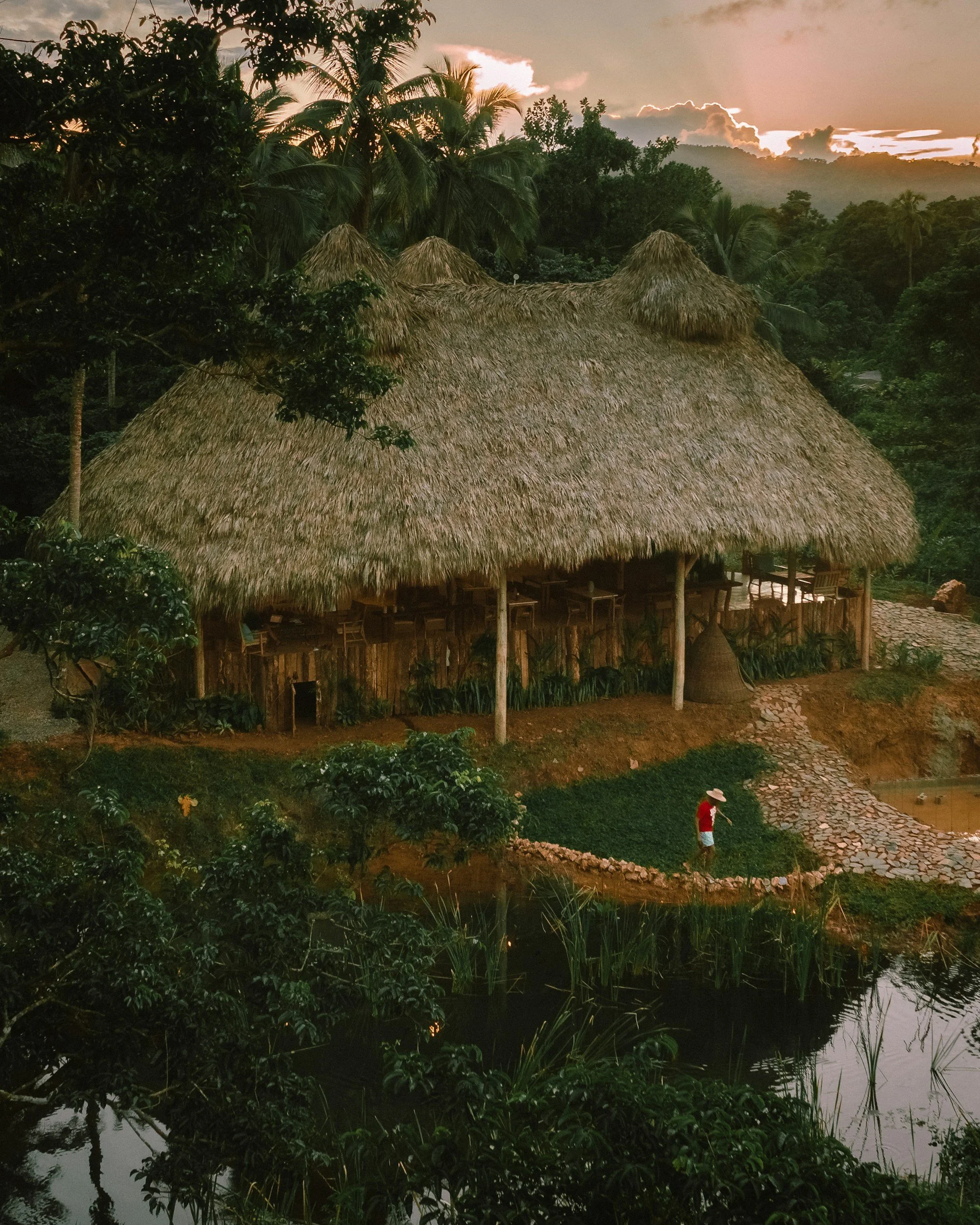 A large thatched-roof hut in a lush tropical setting with palm trees, a pond, and a person in a red shirt and wide-brimmed hat walking on a stone pathway at sunset.
