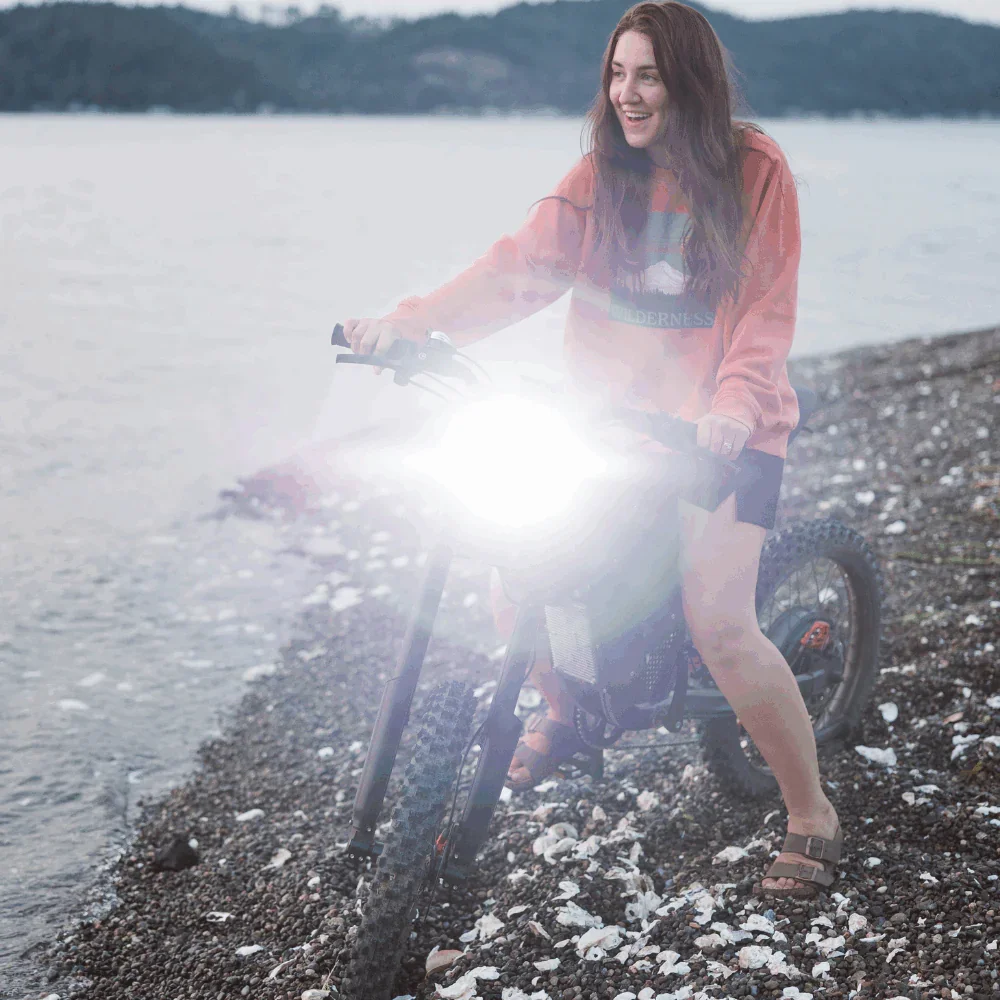 A young woman with long red hair riding a mountain bike on a pebbled shoreline near a body of water, wearing a reddish-orange hoodie, shorts, and sandals, smiling and looking to the side during daytime.