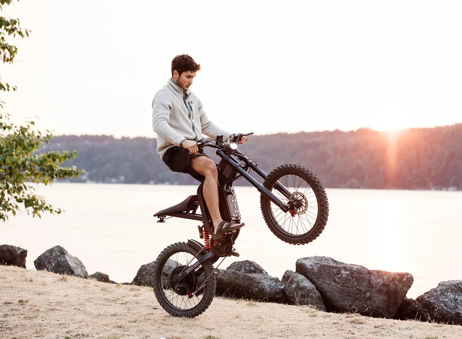 A young man riding an electric dirt bike on rocky terrain near a body of water during sunset.
