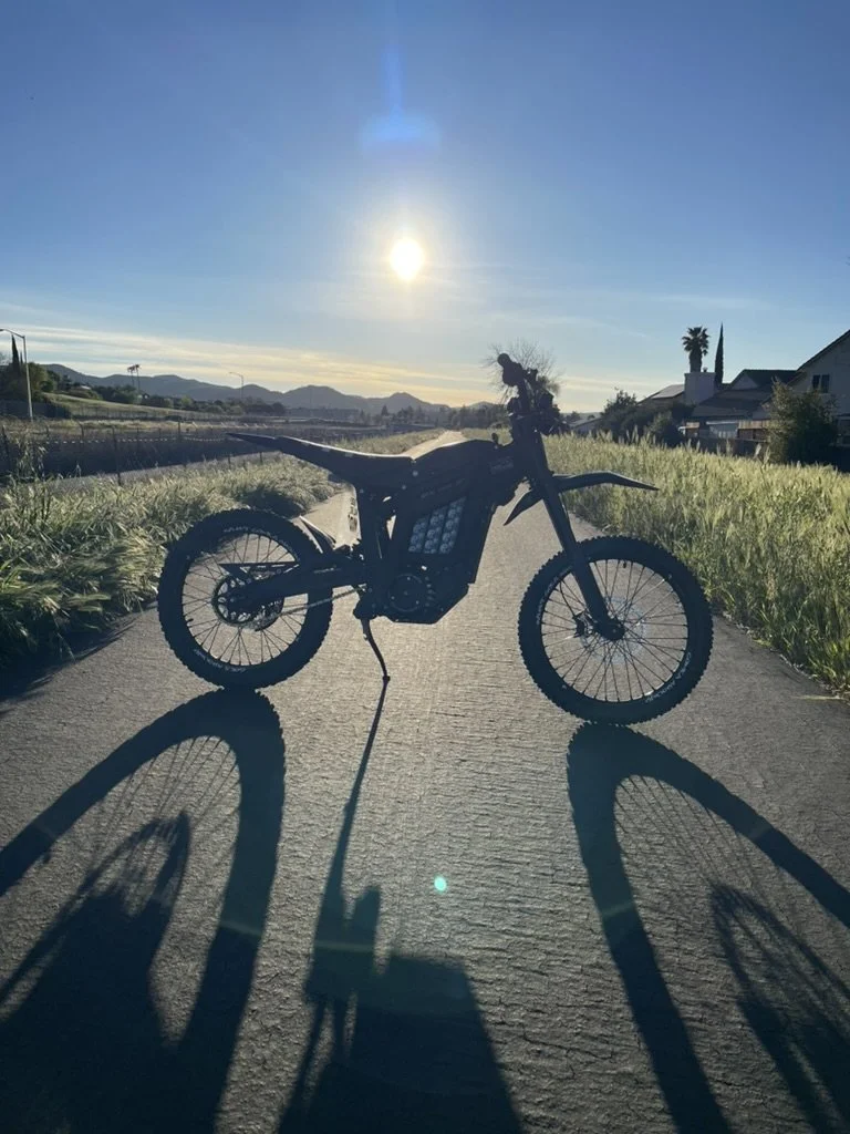 Electric dirt bike parked on a paved road with the sun setting in the background, casting long shadows.