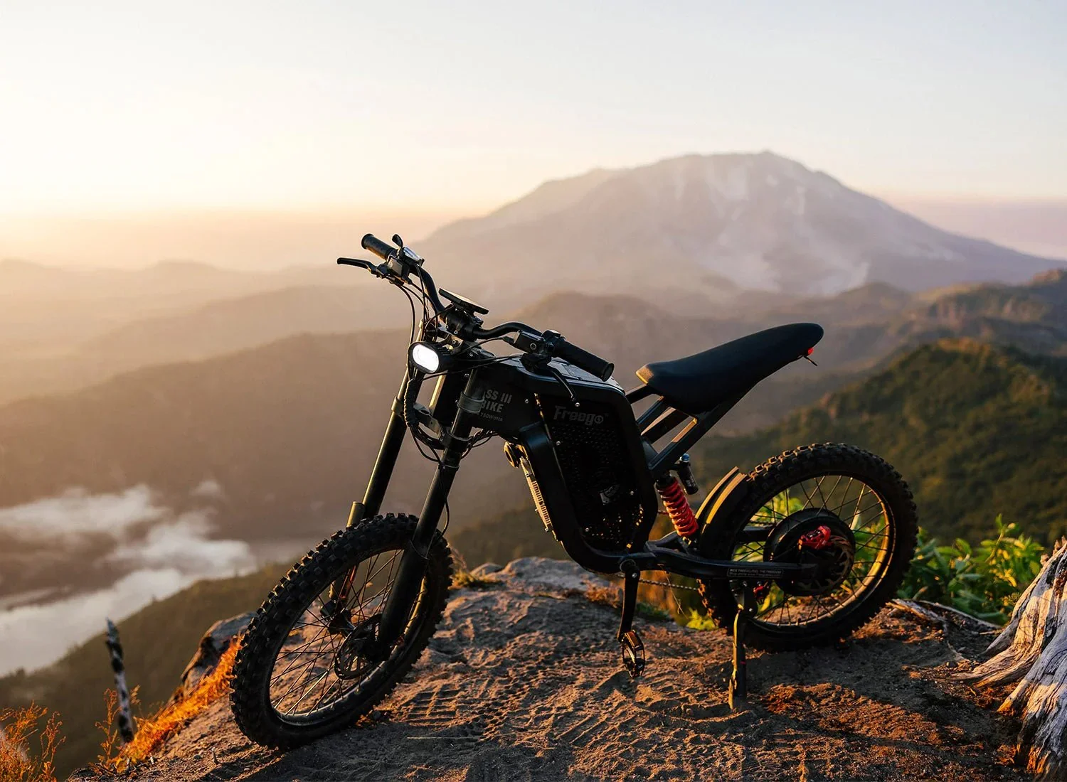 On a rocky peak at sunrise or sunset, an electric mountain bike with thick tires, black frame, and red suspension is positioned with a mountain landscape and distant mountains in the background.