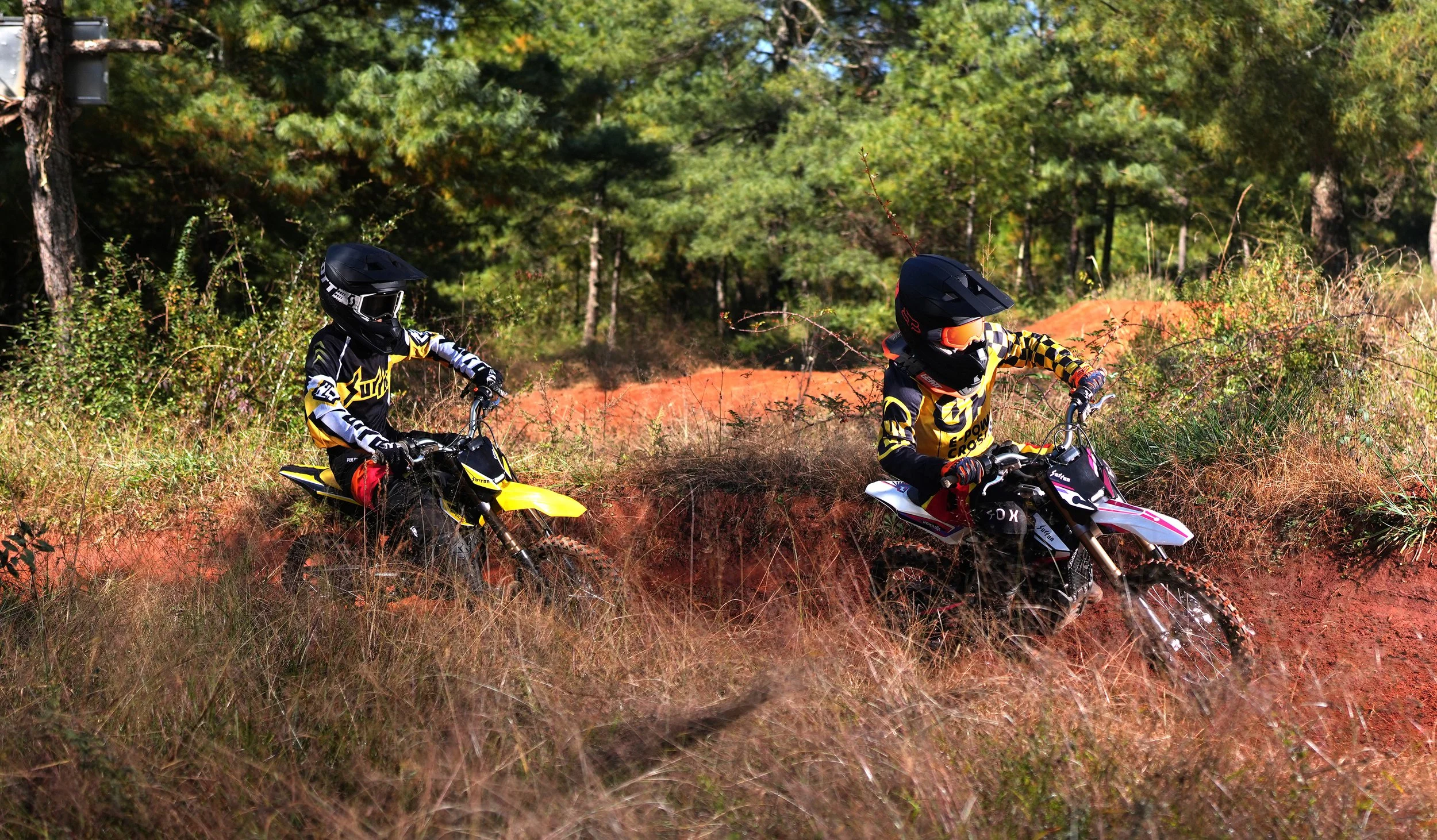 Two children riding dirt bikes through a grassy trail in a forested area, both wearing full helmets and motocross gear.