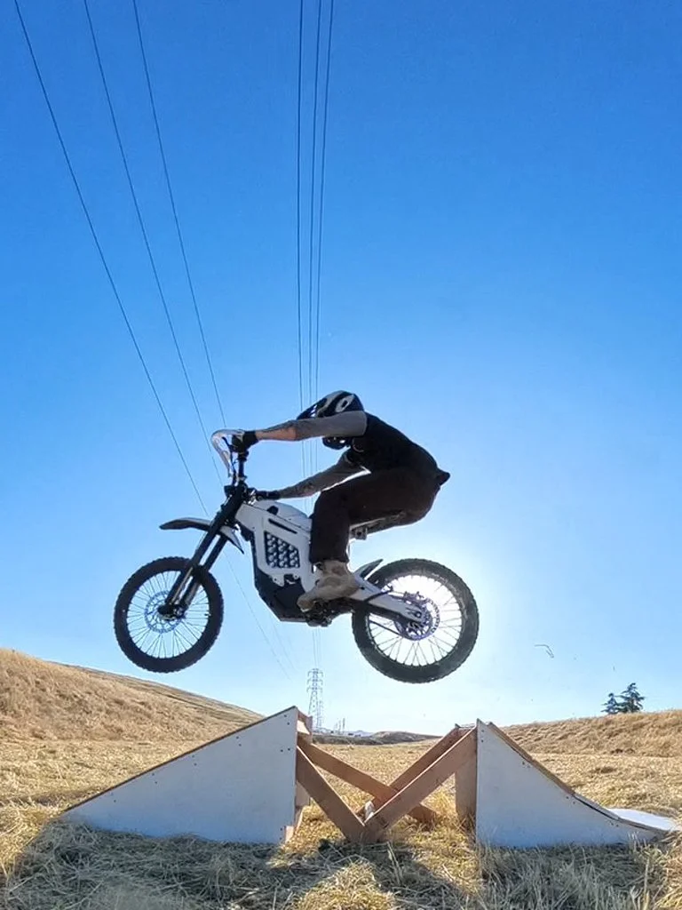 Person wearing a helmet riding an electric bike mid-air over a ramp on a grassy field with power lines in the background.
