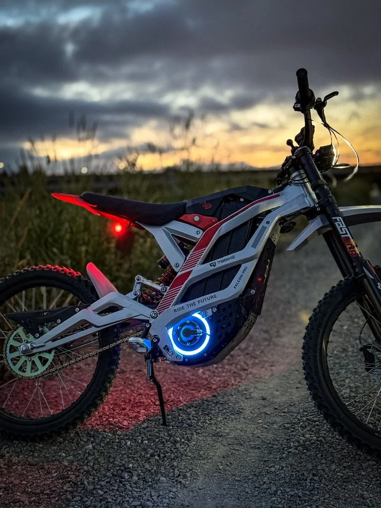 An electric mountain bike parked on a dirt trail at sunset, with a glowing blue circular logo on the battery and illuminated rear light.
