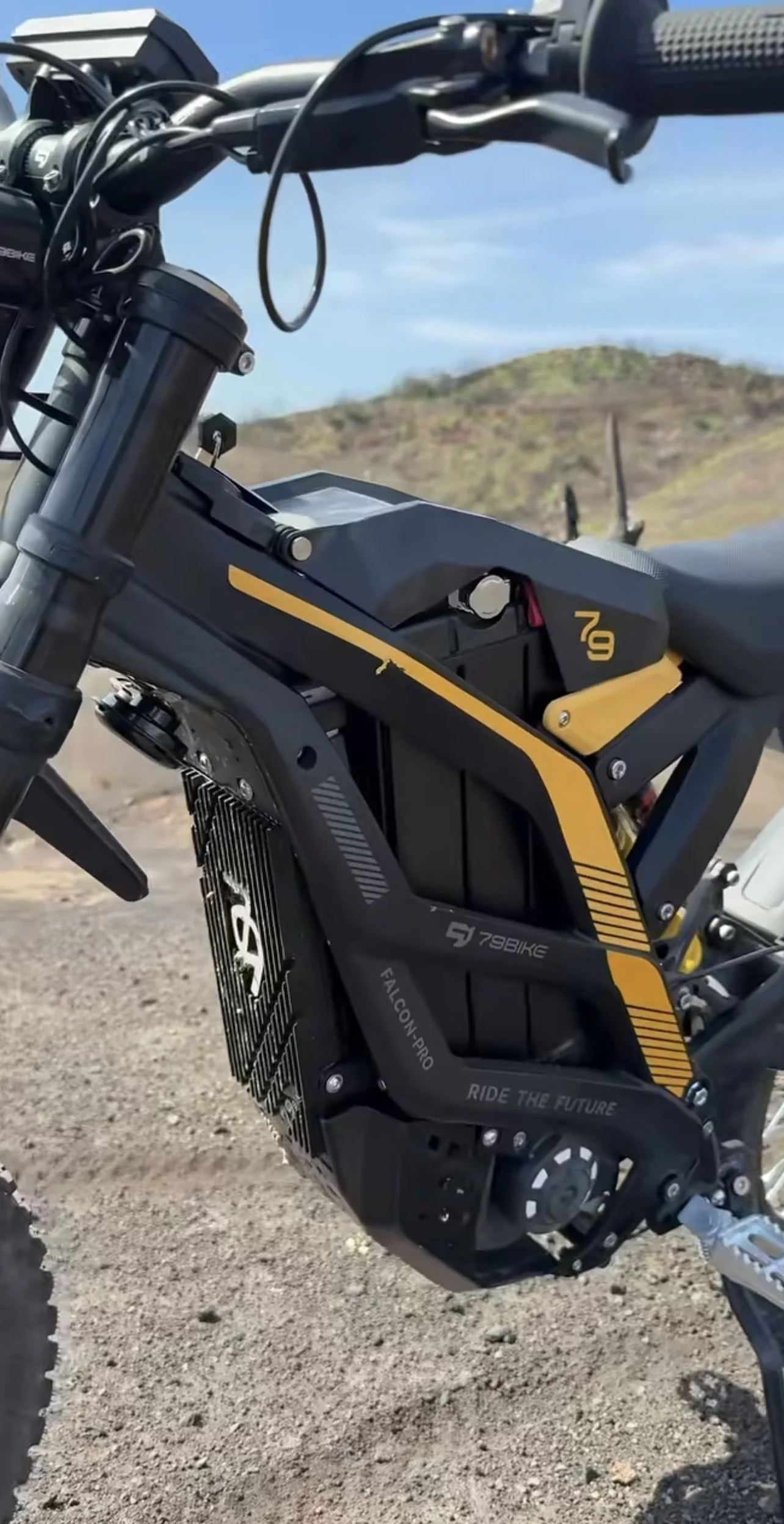 Close-up of an electric dirt bike with a black and yellow design, on a dirt trail with a hilly landscape in the background.
