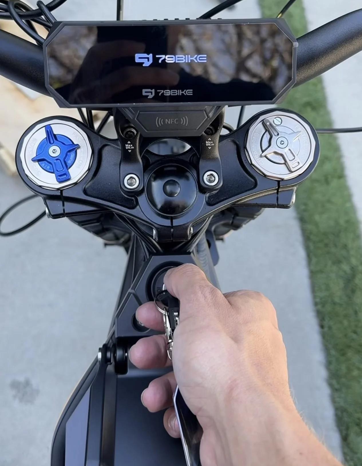 Top view of an electric bike handlebar with digital display, hand holding keys near ignition, on a sidewalk with grass.