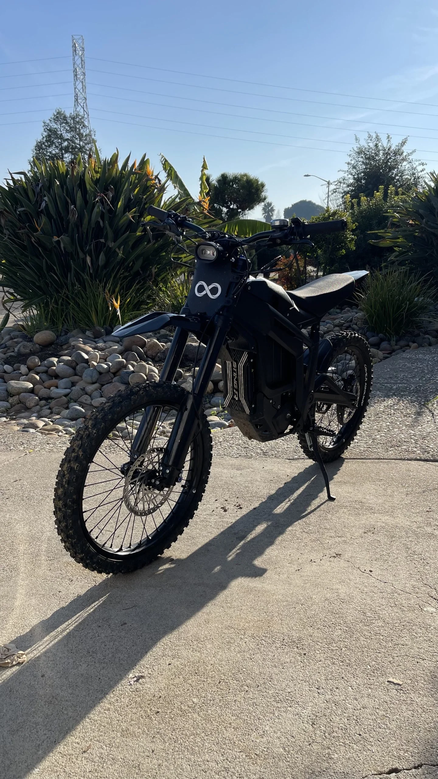 Black electric dirt bike with a white infinity symbol on the front, parked on a concrete surface with desert plants and bushes in the background under a clear blue sky.