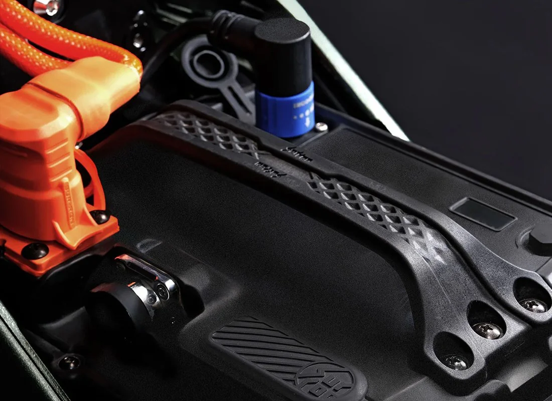 Close-up of a high-performance electric vehicle engine with bright orange cables, a black plastic cover, and a blue component, all mounted on a black chassis.