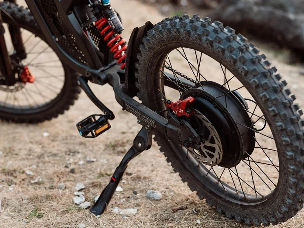 Close-up of an electric mountain bike's rear wheel with thick knobby tire, disc brake, and suspension system on sandy ground.