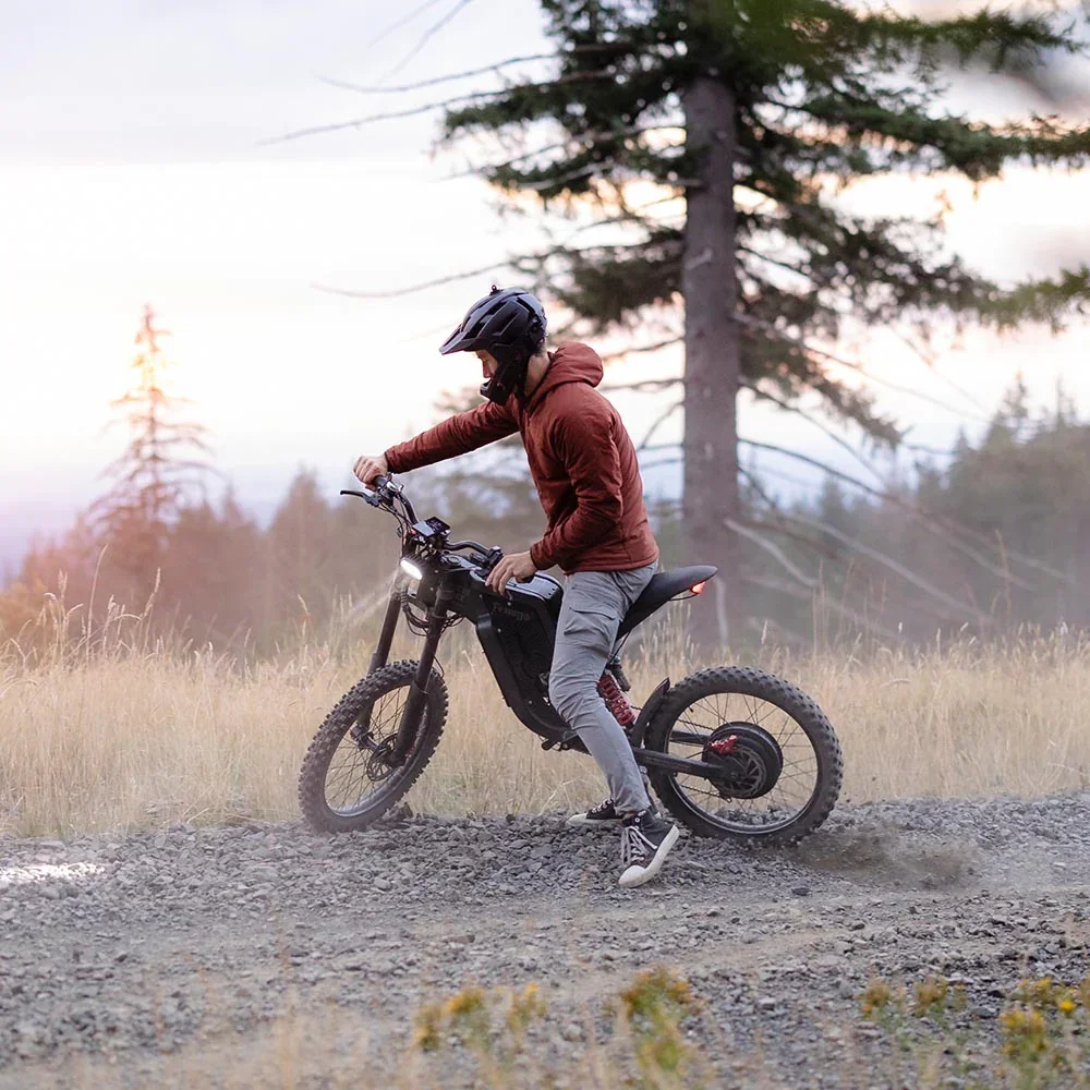Person wearing a helmet and maroon jacket riding a black electric mountain bike on a dirt trail in a natural outdoor setting with trees and grass.